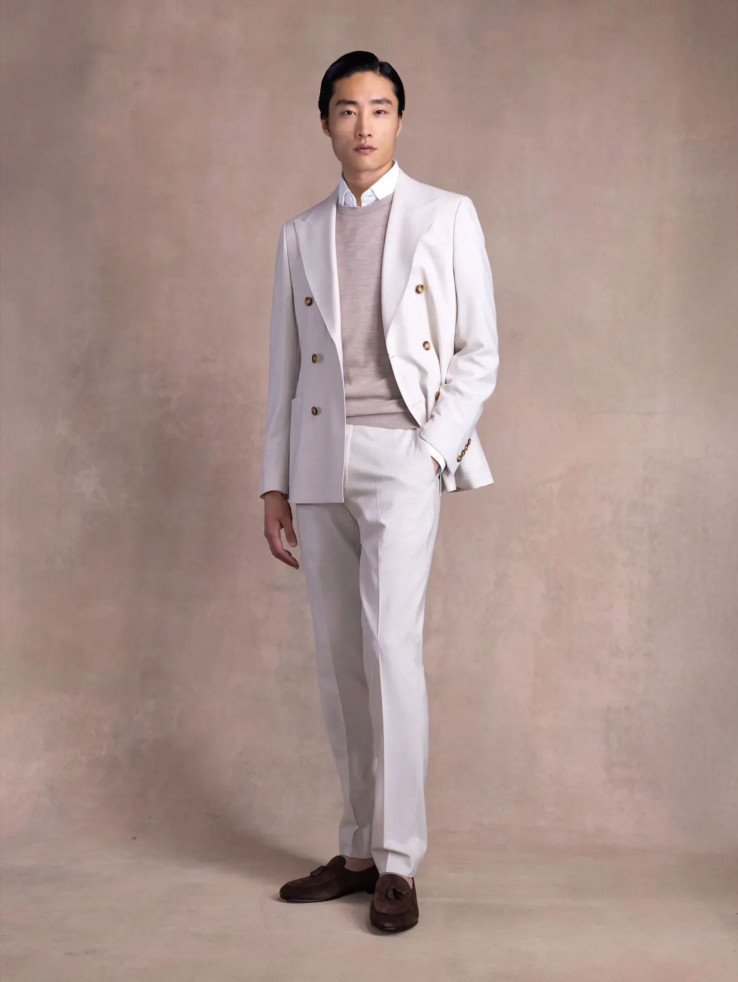 Man with black hair, wearing a white suit jacket and pants, a light beige sweater, white dress shirt, and brown shoes, standing against a plain beige background.