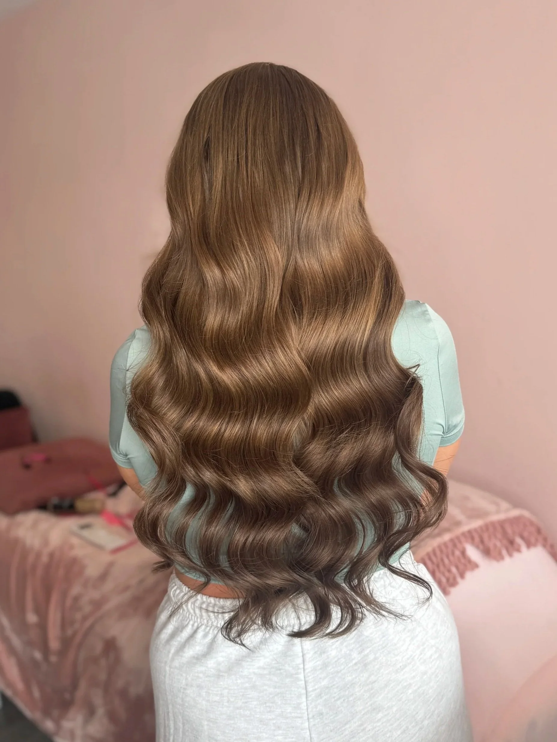 Back of a person with long, wavy, light brown hair wearing a light blue top and white pants, standing in a bedroom with a bed and pink decor.