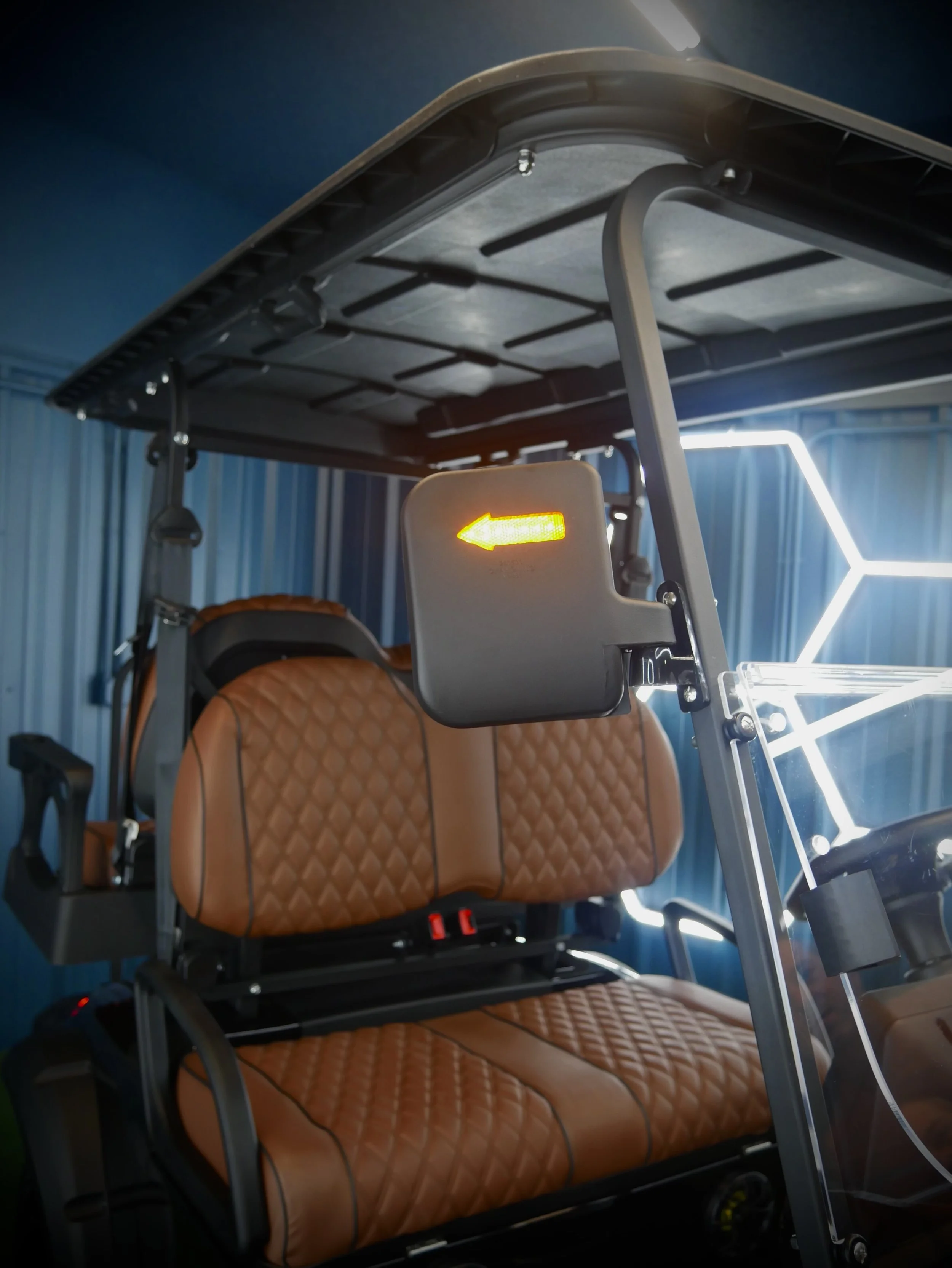 Inside a vehicle, a brown leather seat with a quilted pattern is visible. Behind the seat, there is a headrest and a safety harness. A metal frame and a panel with a yellow arrow illuminated on it are also present, along with a geometric neon light d