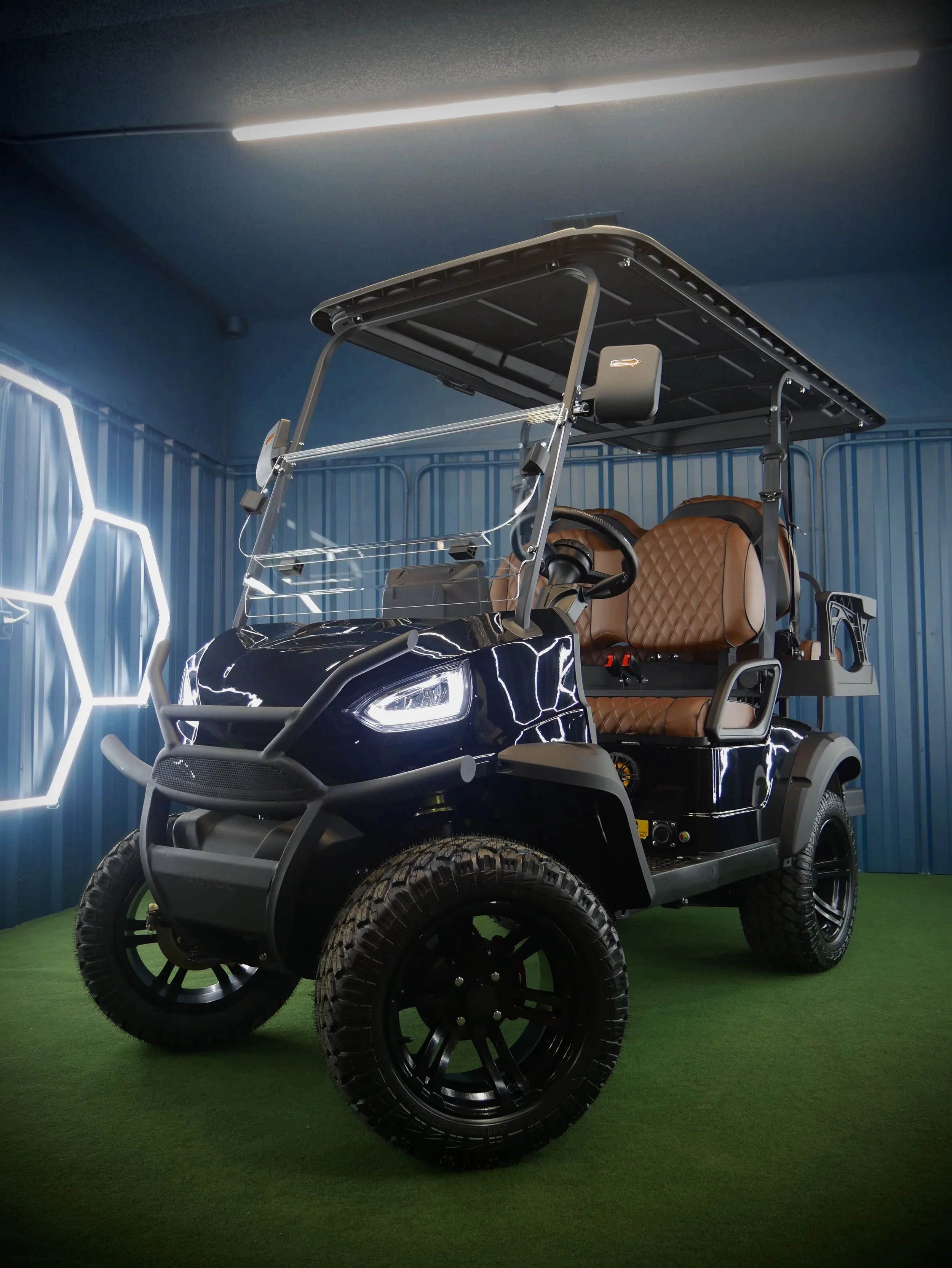 Electric golf cart with tan quilted leather seats, black body, large off-road tires, and a roof, parked on a green turf with a blue wall featuring hexagonal light fixtures in the background.