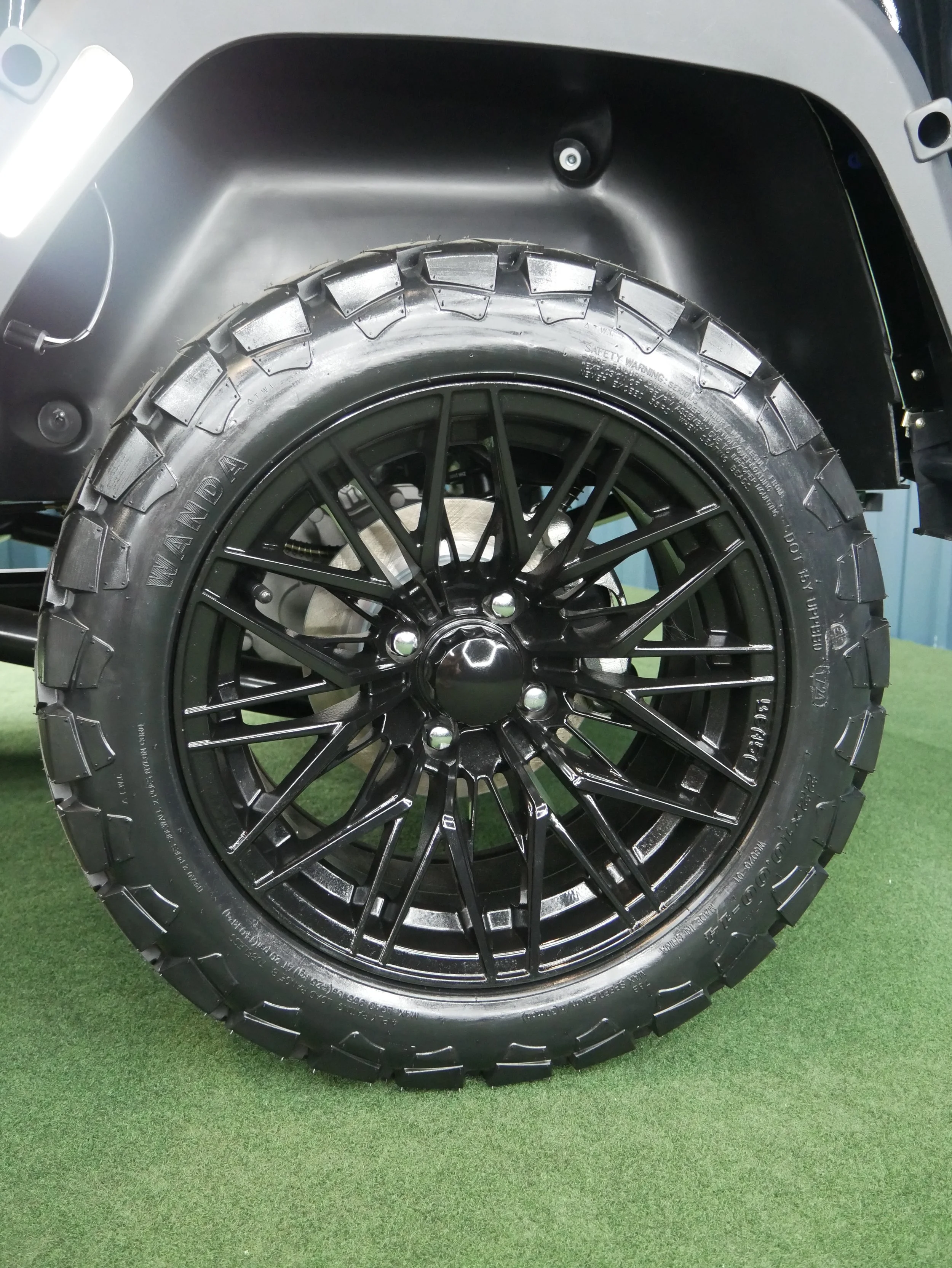Close-up of a black wheel with a rugged tire mounted on a vehicle, set on green artificial grass.