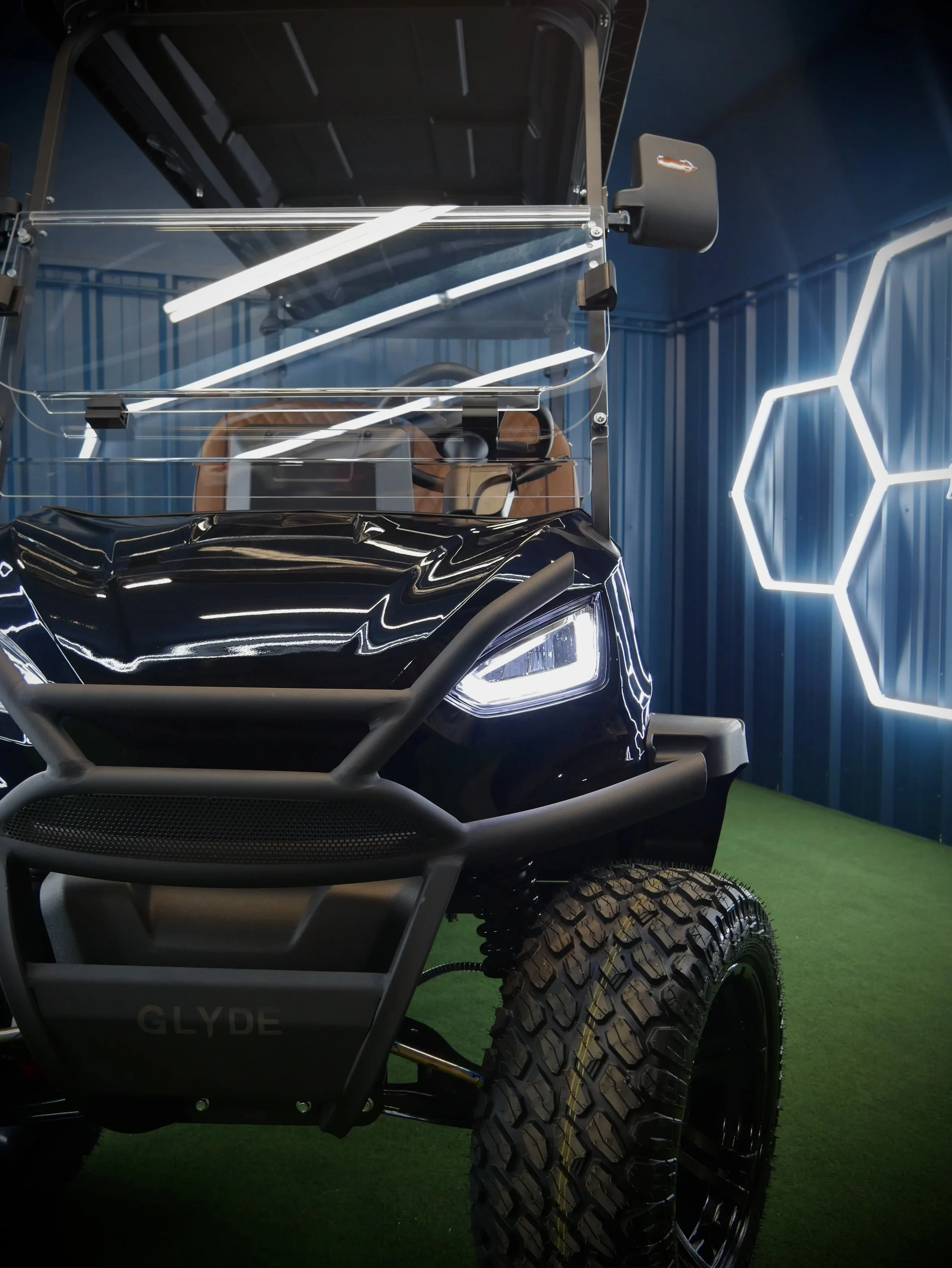 Close-up of an off-road electric vehicle with rugged tires, a black body, and decorative white LED strip lights on a green artificial turf floor, with a modern geometric LED wall design in the background.