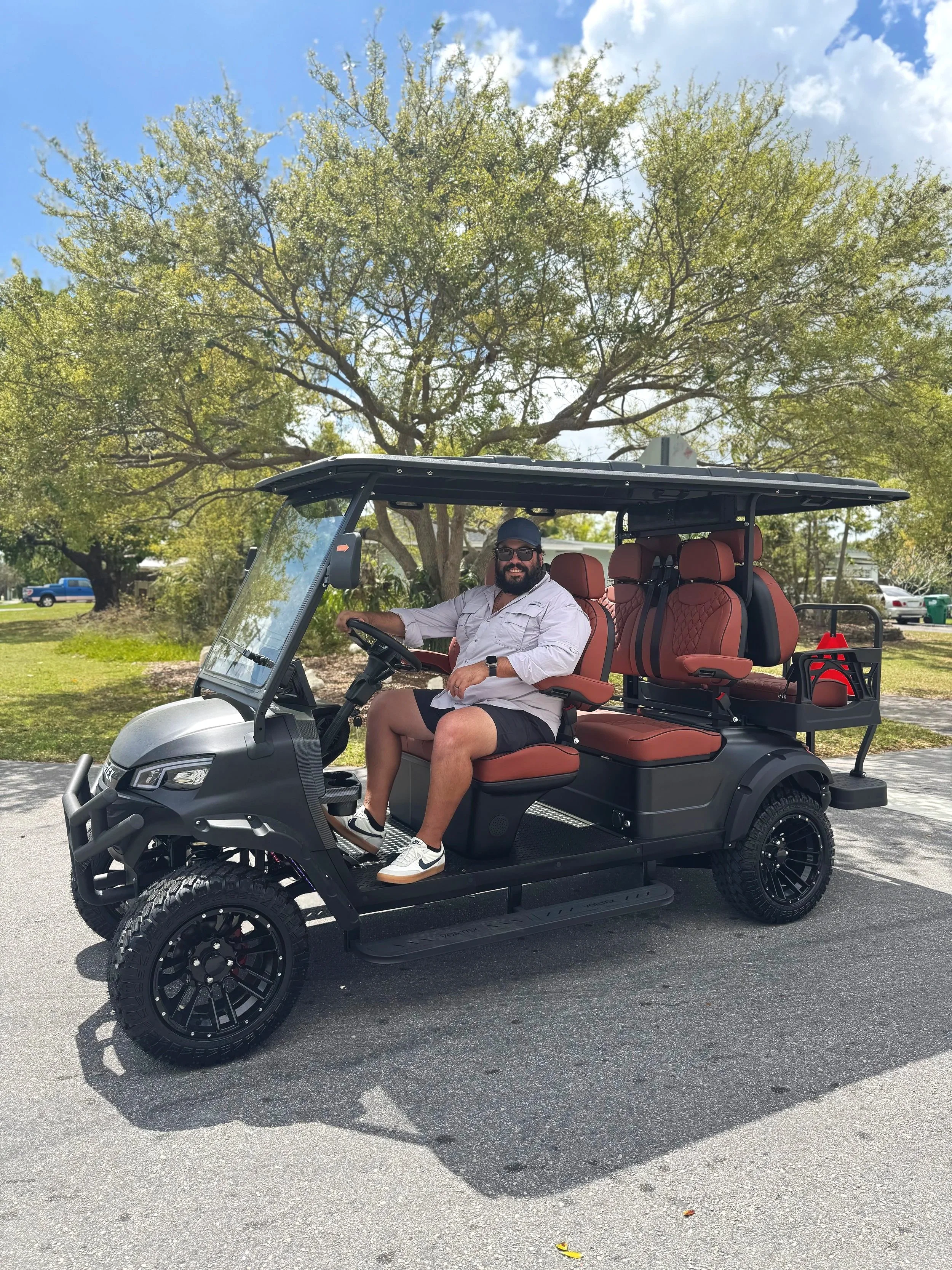 A man with a beard, sunglasses, and a gray cap sitting in a black electric golf cart with brown seats, parked outdoors under a tree on a sunny day.