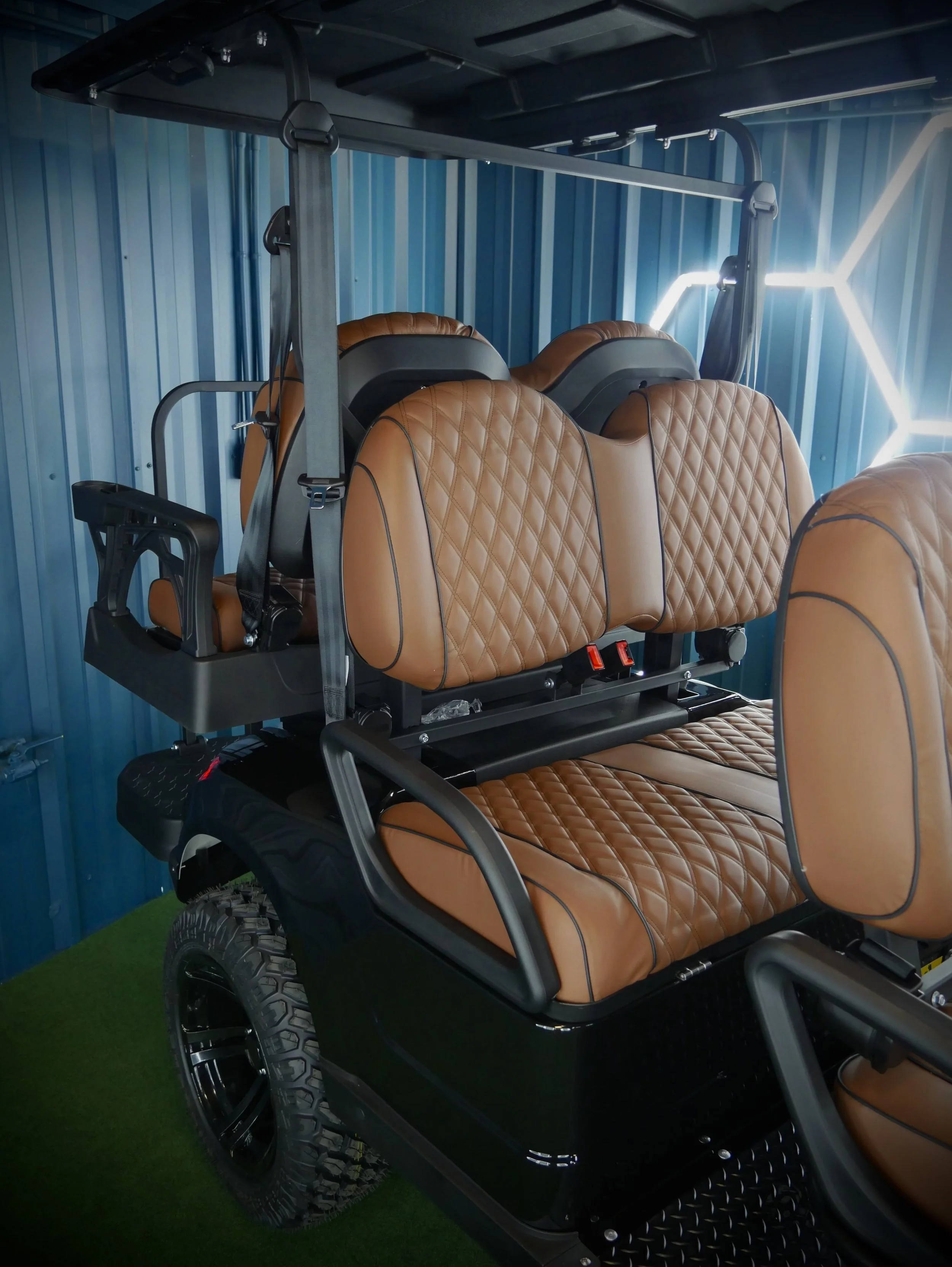 All-terrain golf cart with tan leather seats and a black frame, parked on green artificial grass against a blue wall with neon lighting accents.