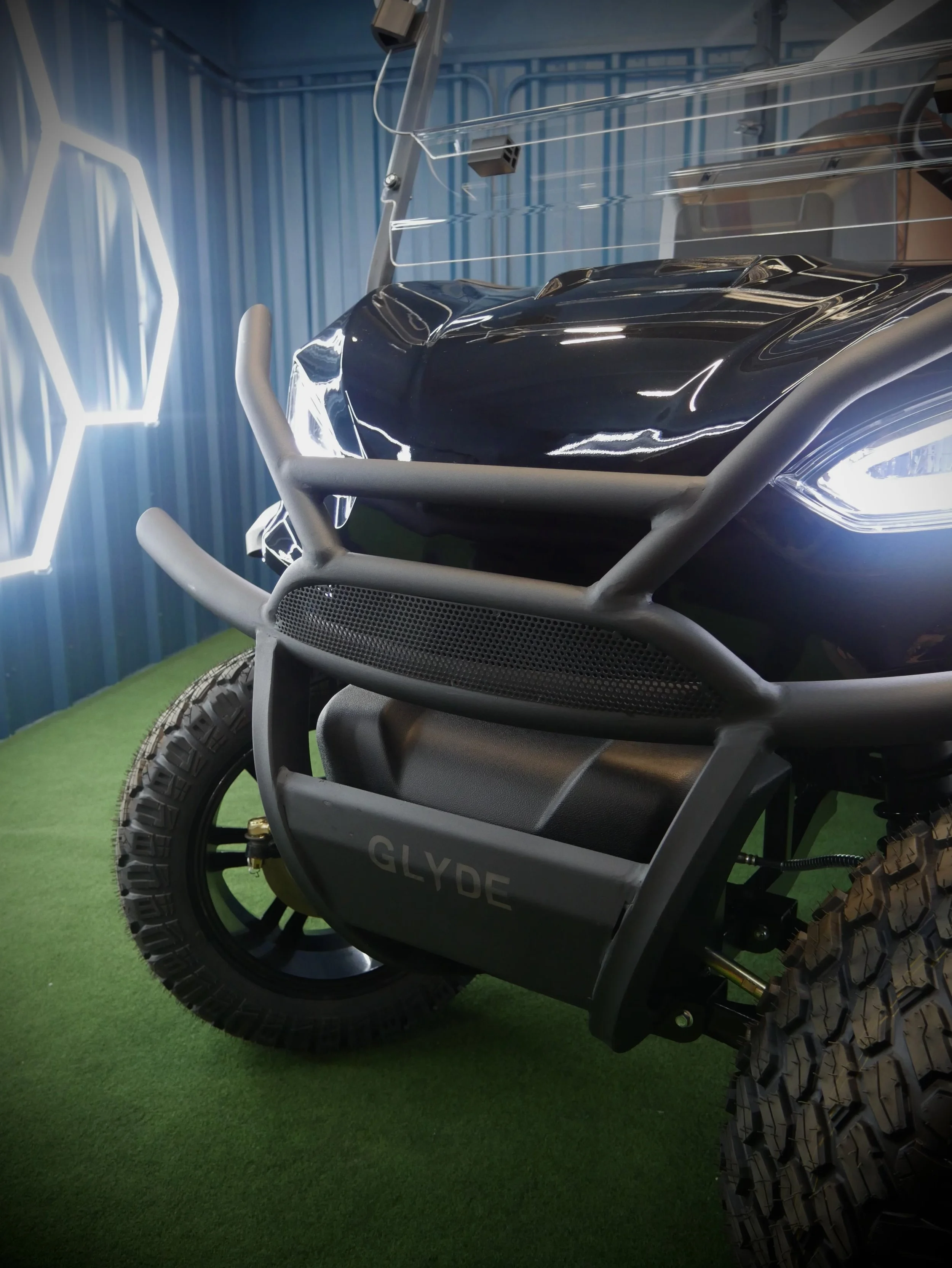 Close-up view of an off-road electric scooter with black body, rugged tires, and a sturdy frame, displayed on a green floor in a room with blue walls.