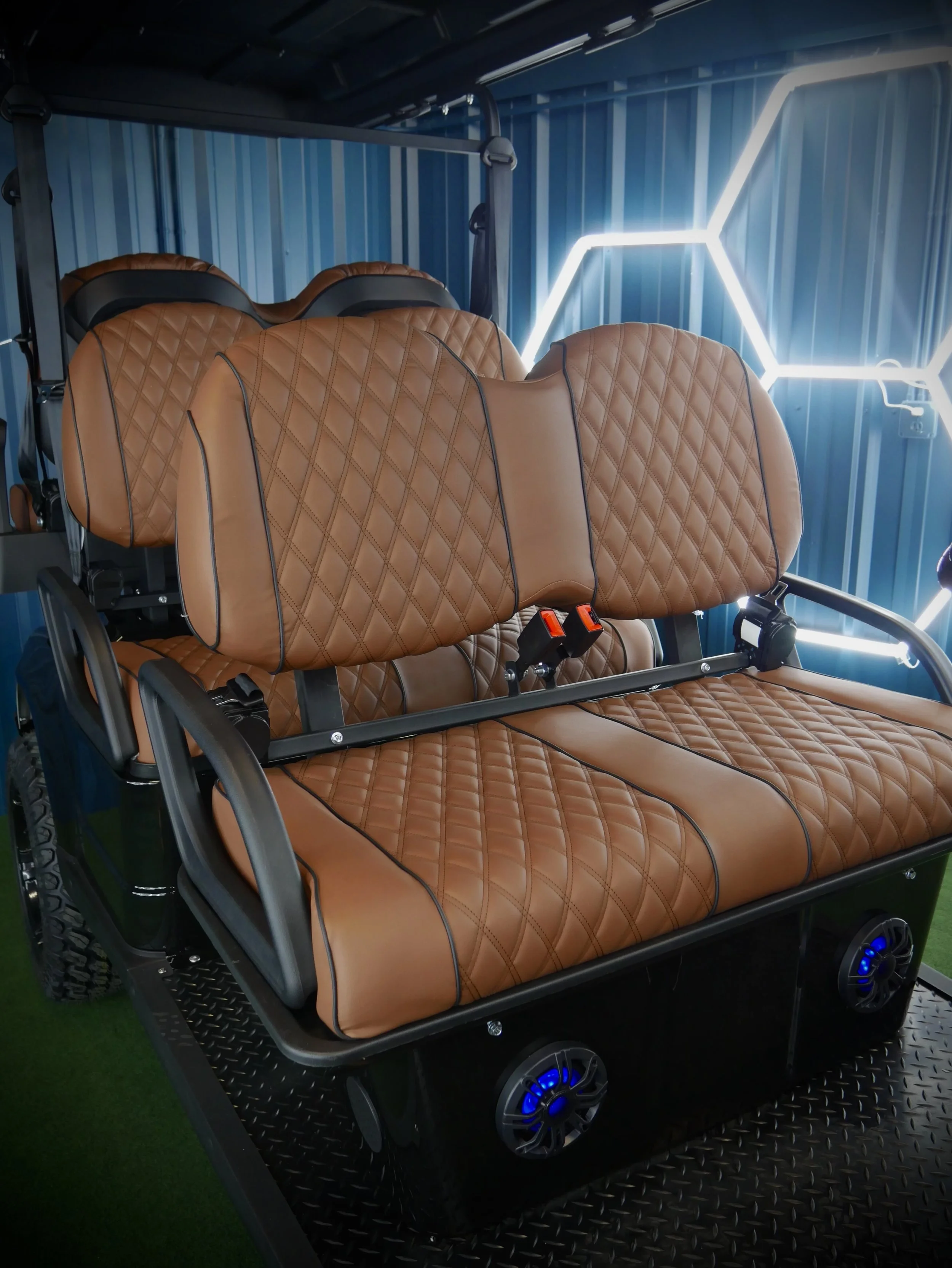 A golf cart with tan quilted leather seats on a black metal platform with blue wheels and neon light hexagon designs on the wall behind it.