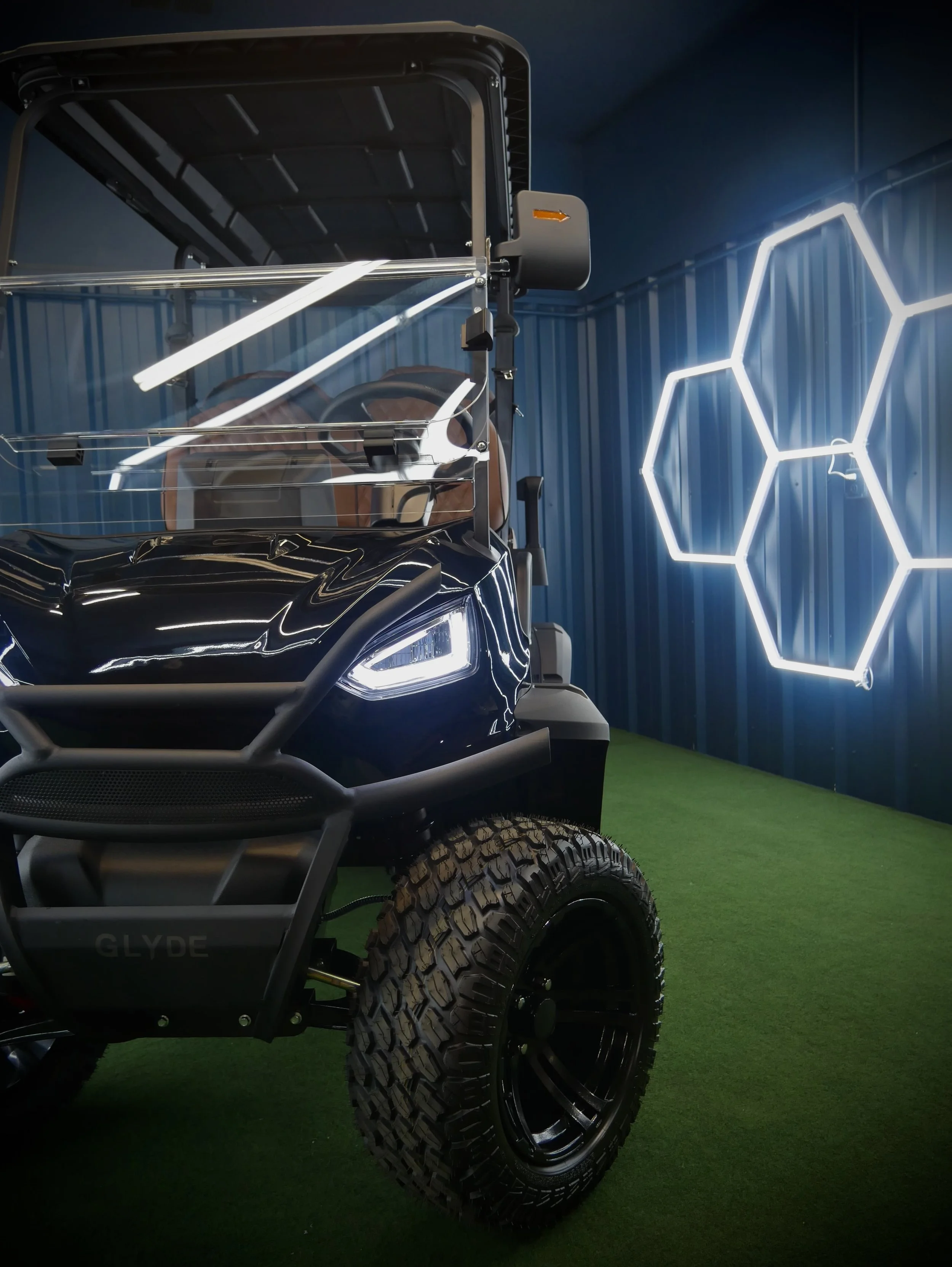 Close-up of a futuristic black off-road vehicle with large tires, a transparent windshield, and illuminated hexagonal wall design in the background.