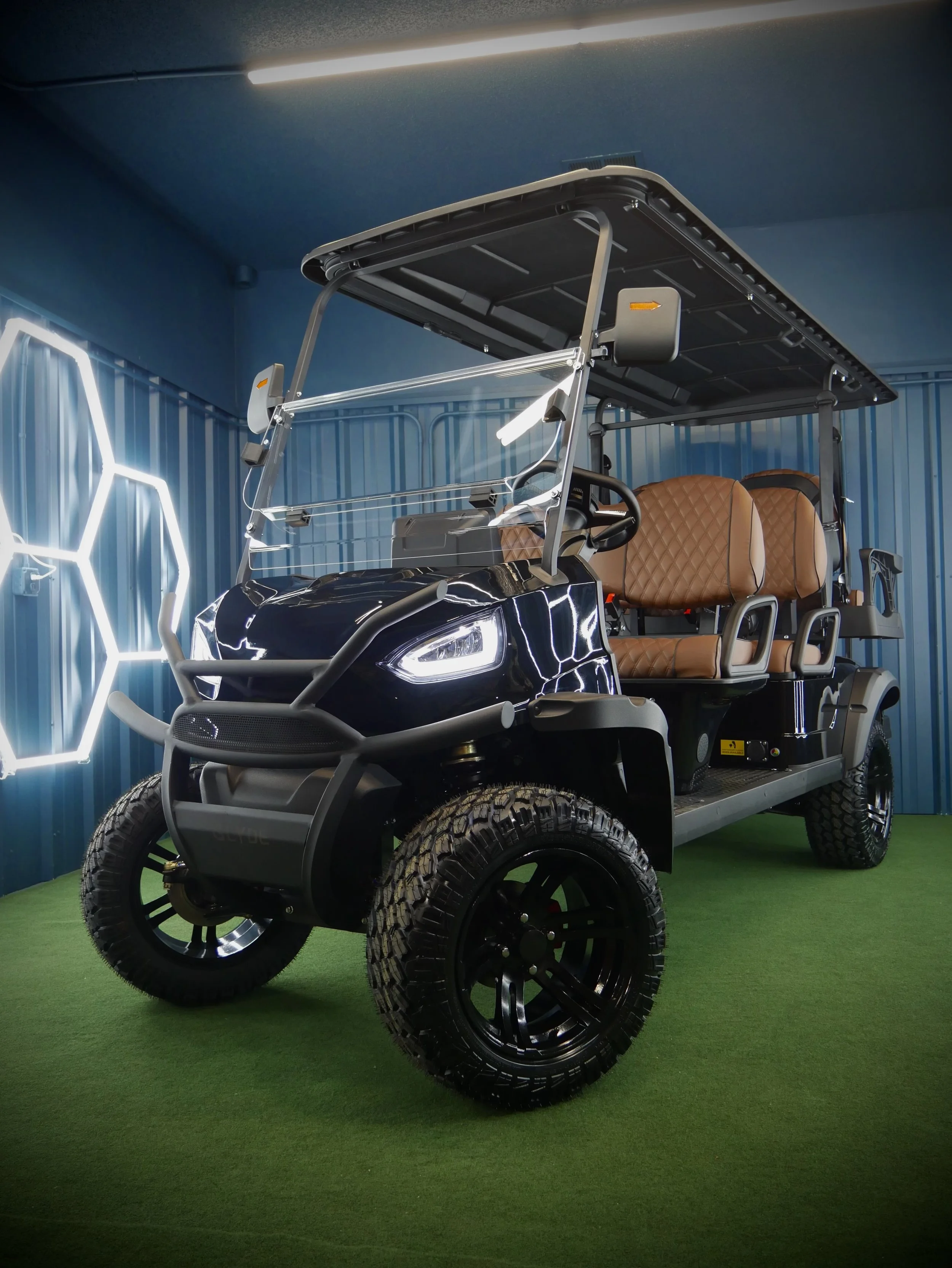 A black golf cart with tan leather seats and a roof is displayed indoors. The walls are blue with white hexagonal lighting accents, and the floor is green, resembling grass.
