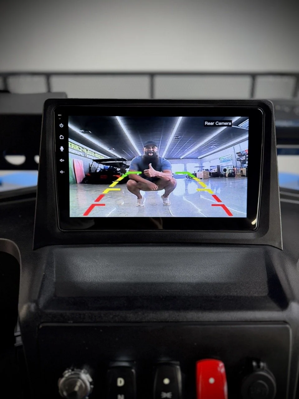 Car interior display showing a rearview camera image of a person squatting in a gym, giving a thumbs-up. The screen has colored guide lines to assist with reversing.