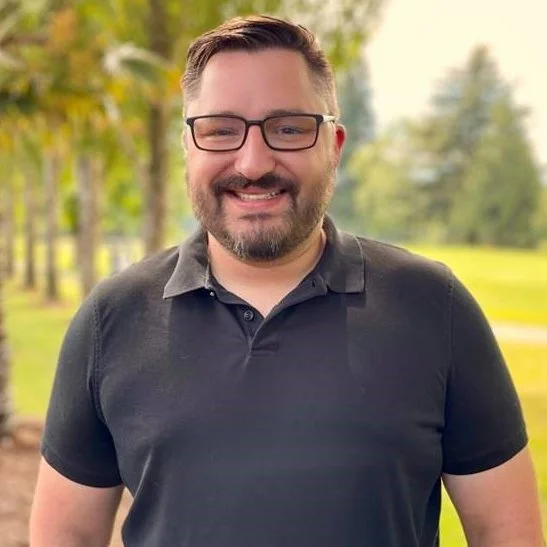 Jordan, the owner and CEO of J3 Consulting, with glasses and beard outdoors on sunny day, trees and grass in background.
