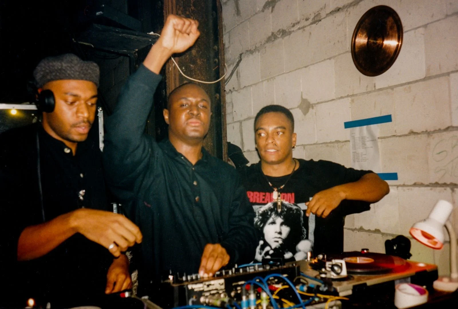 Three young men in a room with a concrete wall; one in the middle raising his fist, one on the right wearing a black shirt with a woman's face on it, and the third on the left with headphones. There is DJ equipment and a small lamp on the table.