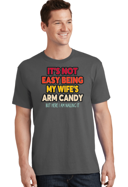 A smiling man wearing a dark gray T-shirt with colorful text that reads, "It's not easy being my wife's arm candy but here I am nailing it."