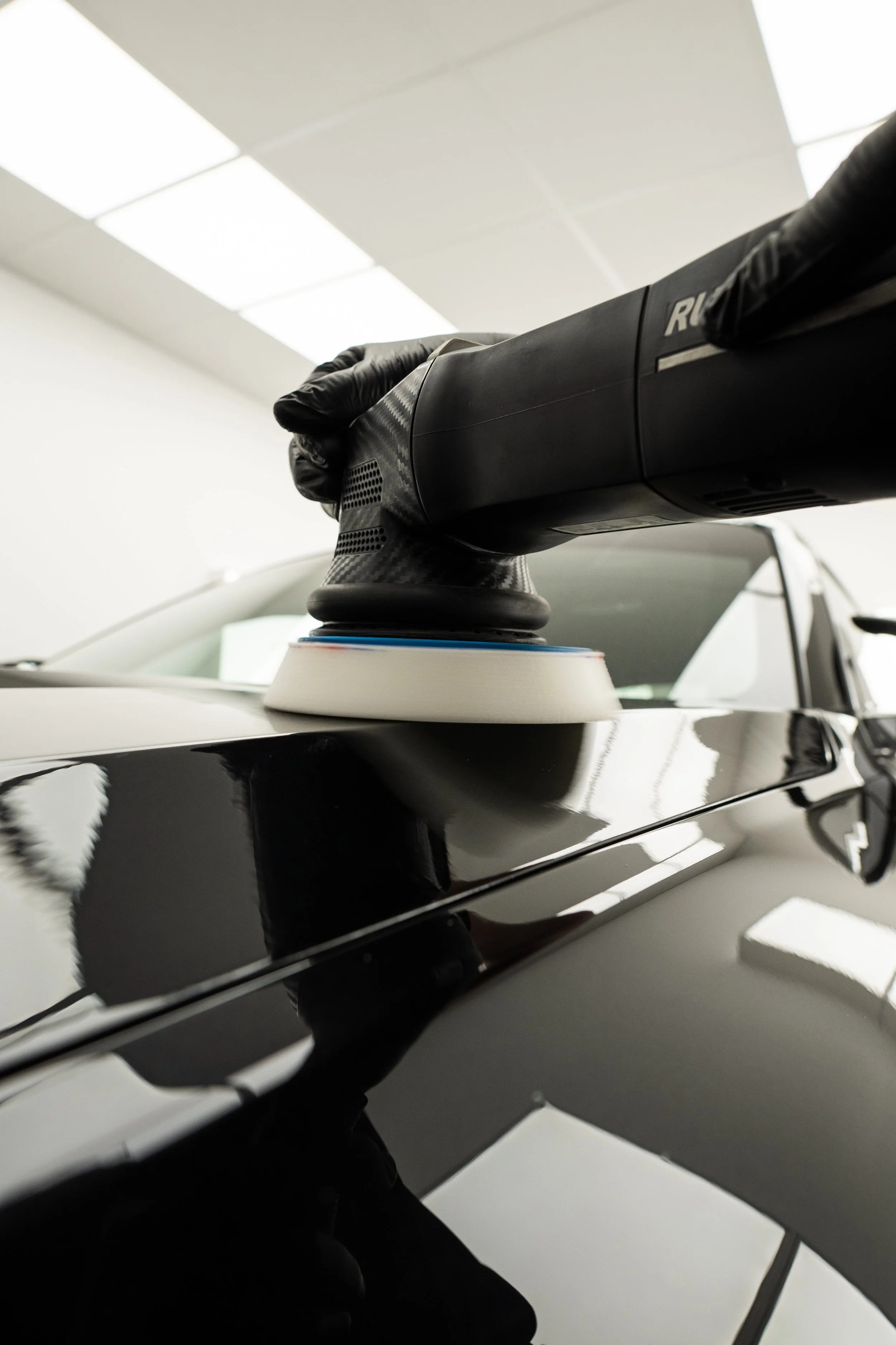 Person wearing black gloves polishing a black car with a polisher in an indoor space.