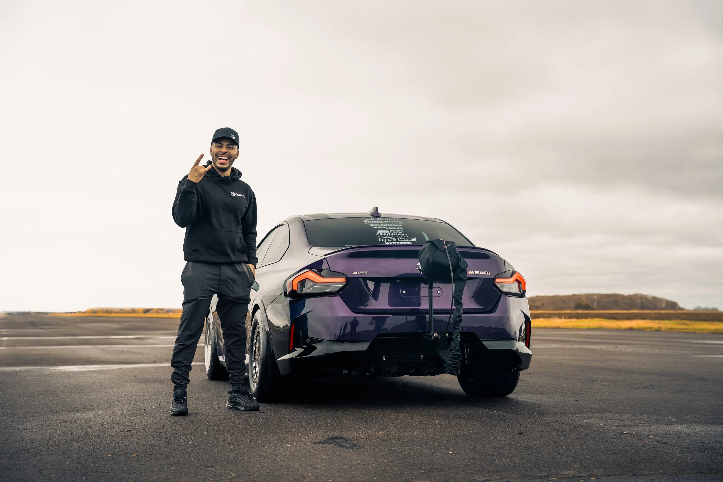 A man stands next to a purple electric car on an open road, smiling and making a rock and roll hand gesture.