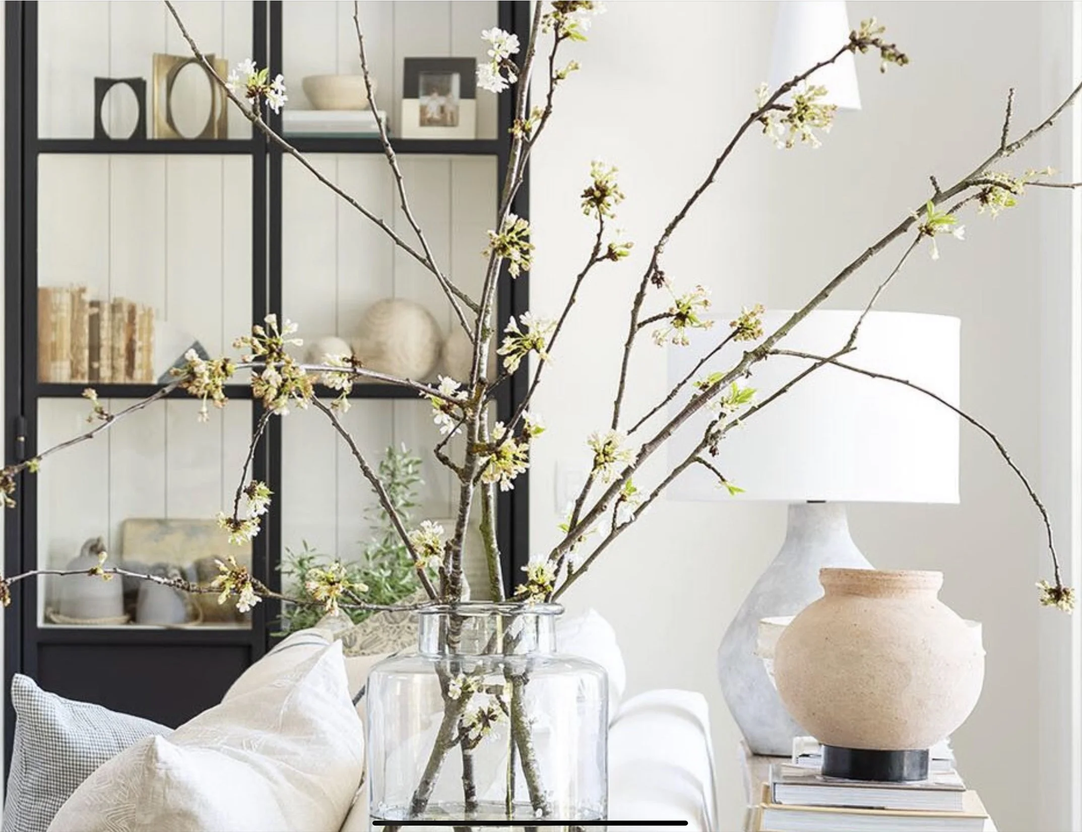A clear glass vase with brown branches and small white blossoms on a white sofa. A beige ceramic lamp and a stack of books are in the background.