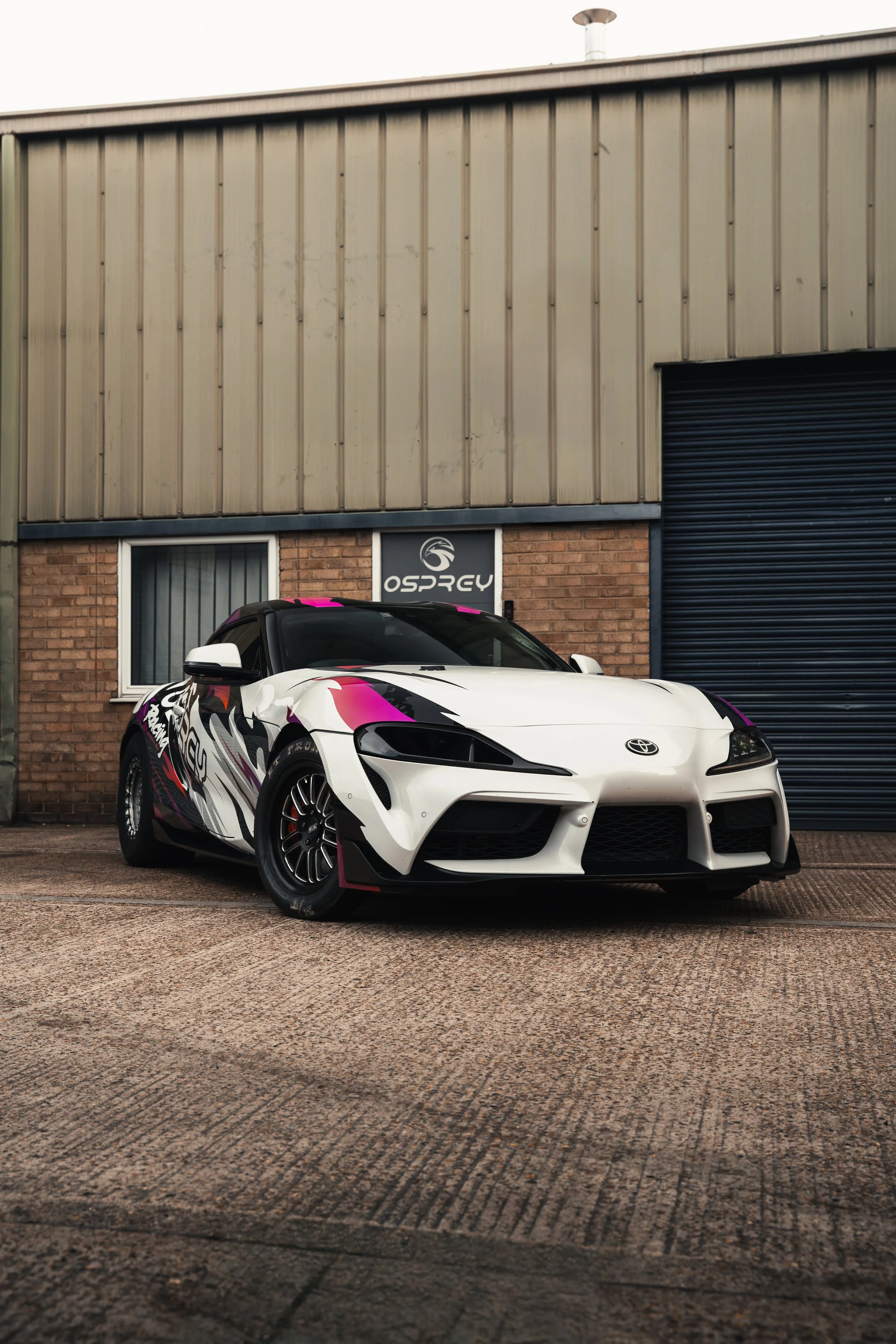 A white sports car with black, pink, and purple graphics parked in front of a brick building with a sign reading 'OSPREY' and a closed black roller door.