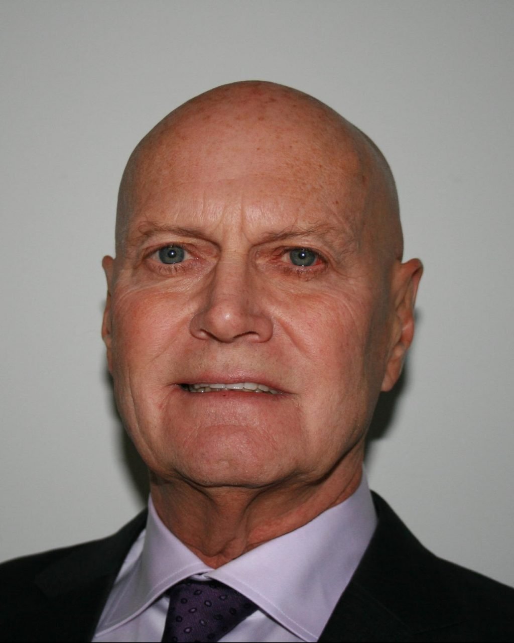 Close-up of an older man with a bald head, blue eyes, wearing a suit and tie, against a plain background.