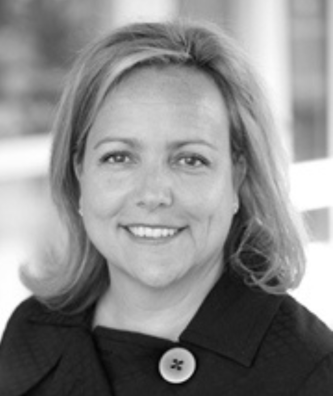 Black and white photo of a smiling woman with shoulder-length wavy hair, wearing a dark coat with a large button.