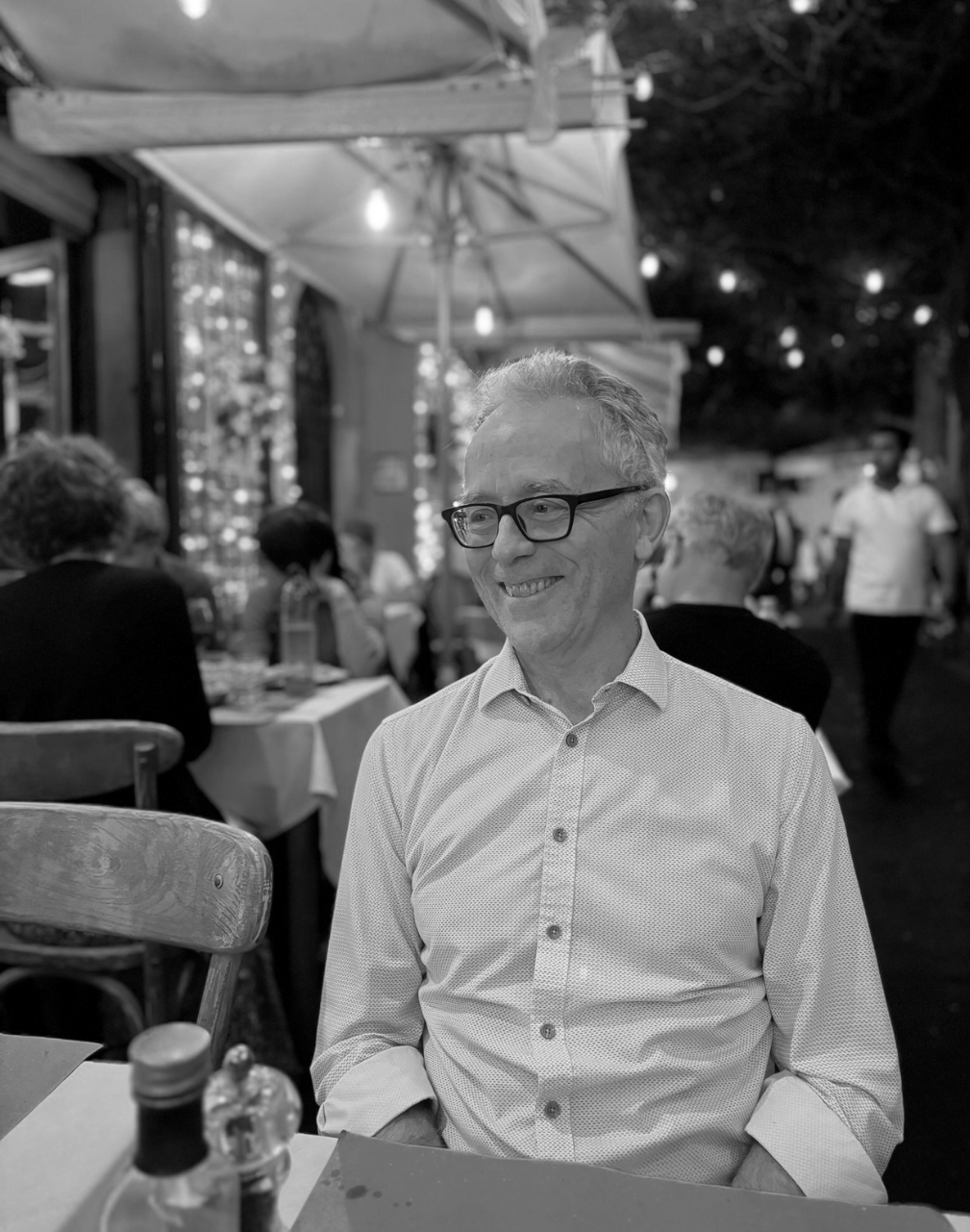 A man with glasses smiling at a restaurant or outdoor dining setting during the evening, with tables, chairs, and other people in the background.