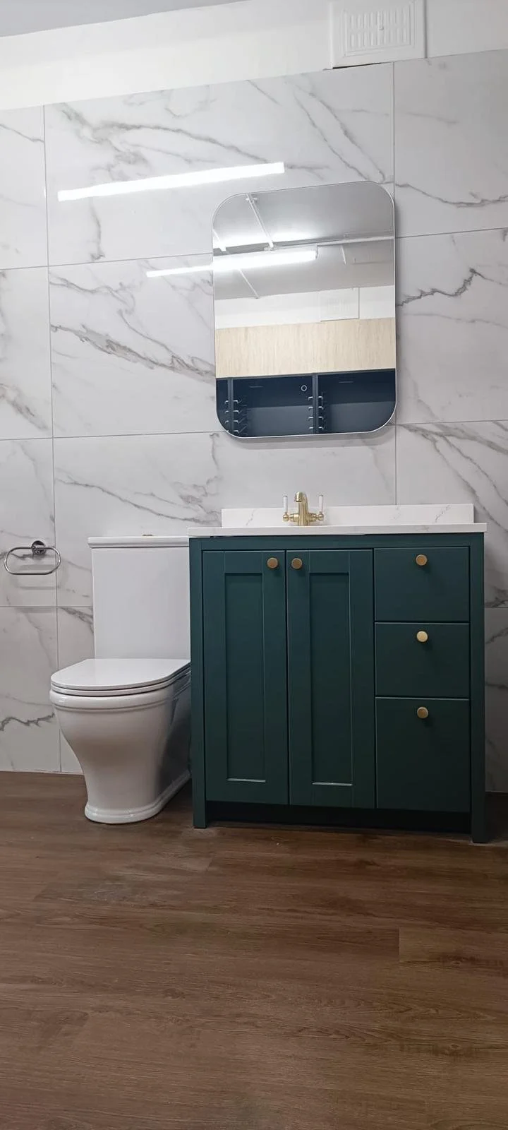 Modern bathroom with marble tiled wall, white toilet, teal vanity with gold accents, small white marble countertop, and oval mirror.