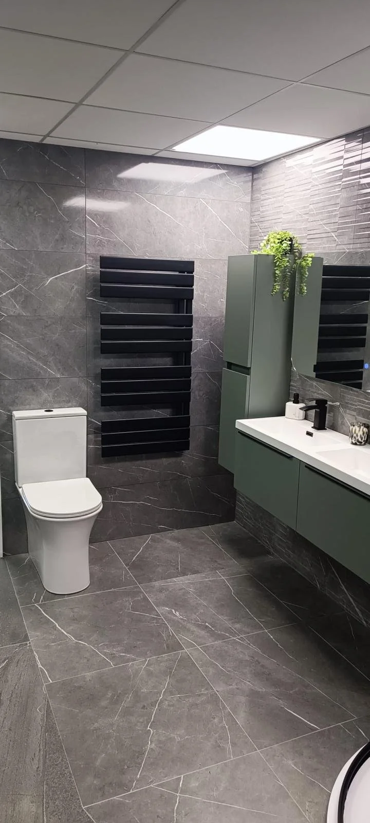 Modern bathroom with a white toilet, black wall-mounted radiator, green cabinetry, and marble-look tiles