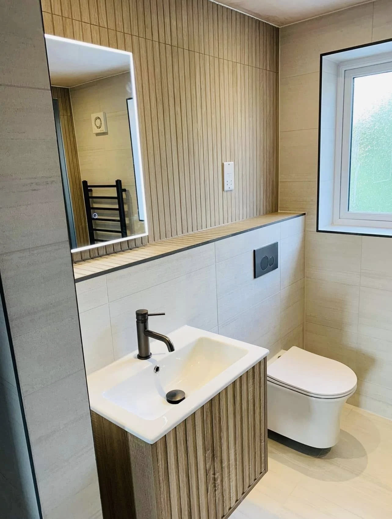 Modern bathroom with wooden panel walls, a small white sink with a black faucet, a mirror with backlight, a wall-mounted toilet, a window with frosted glass, and a black towel rack reflected in the mirror.