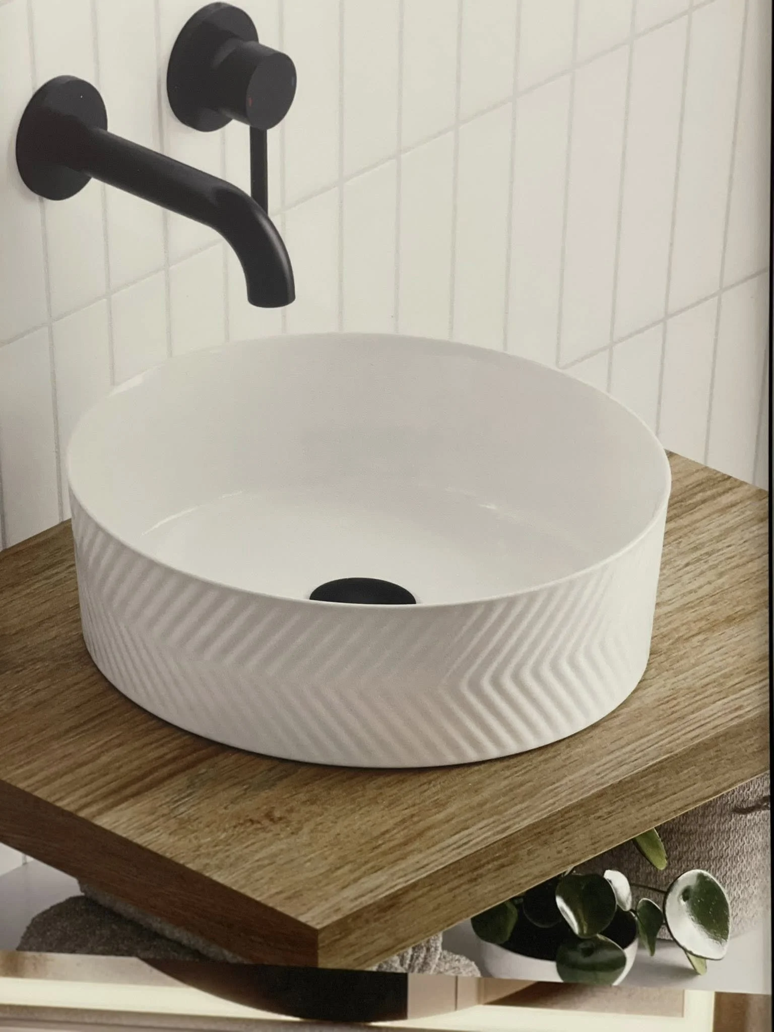 A white ceramic vessel sink with a textured pattern on its exterior, mounted on a wooden countertop. There is a black wall-mounted faucet above the sink and white tiles on the wall behind it.