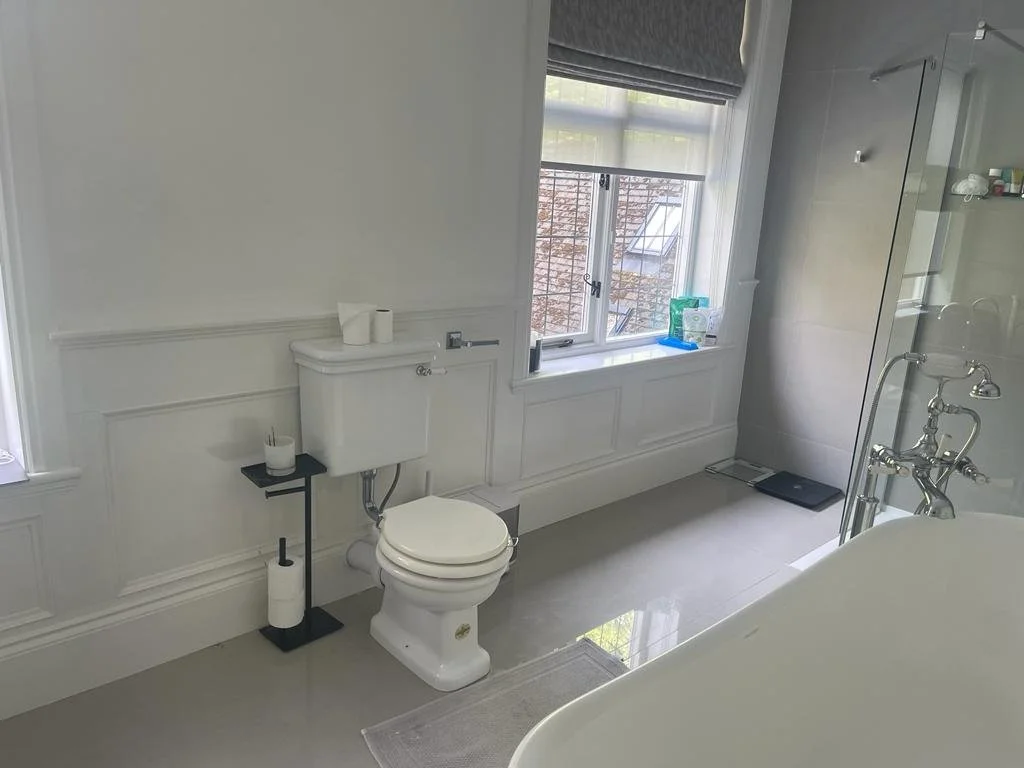Bright, white bathroom with a toilet, a bathtub, a large window, and a glass shower enclosure.