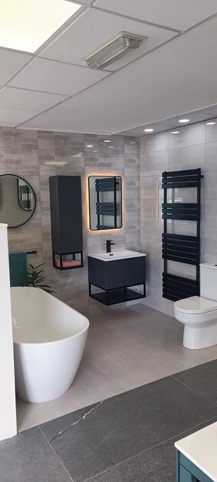 Modern bathroom with a white freestanding bathtub, a floating vanity with a black sink and mirror, black heated towel racks, a toilet, and gray tiled walls and floor.