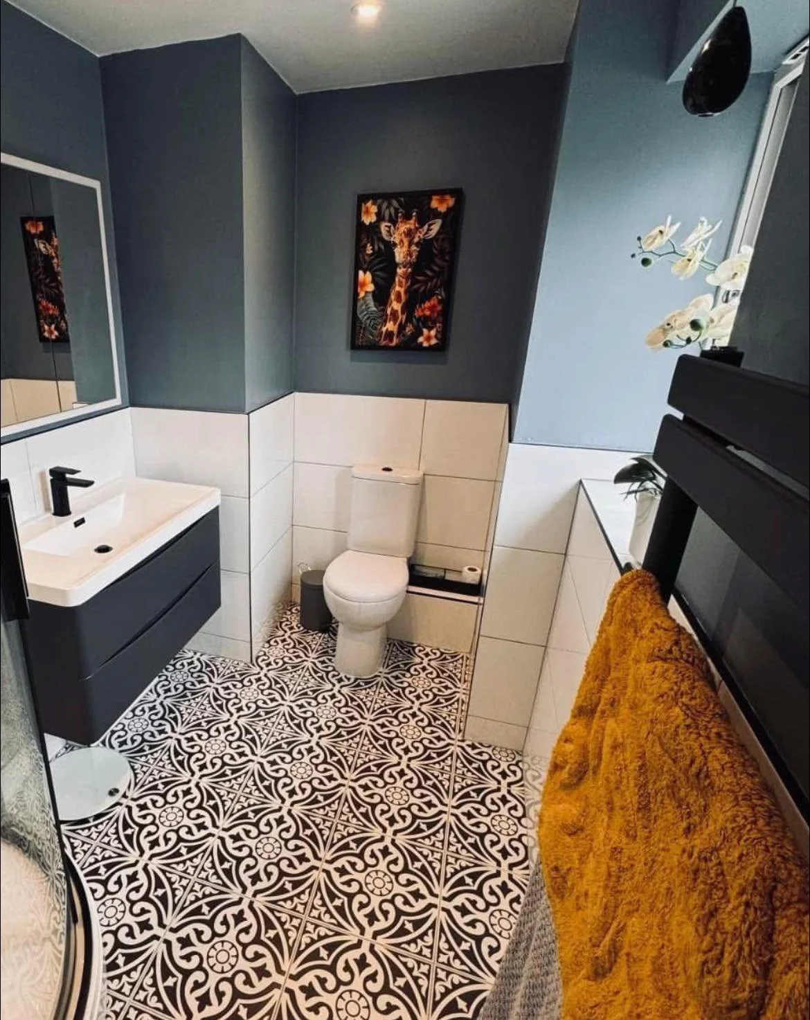 A modern bathroom with a dark blue wall, a white toilet, a black sink vanity, patterned black and white floor tiles, a mirror, a framed giraffe and floral art, a towel rack with yellow towels, and a window with white orchids.