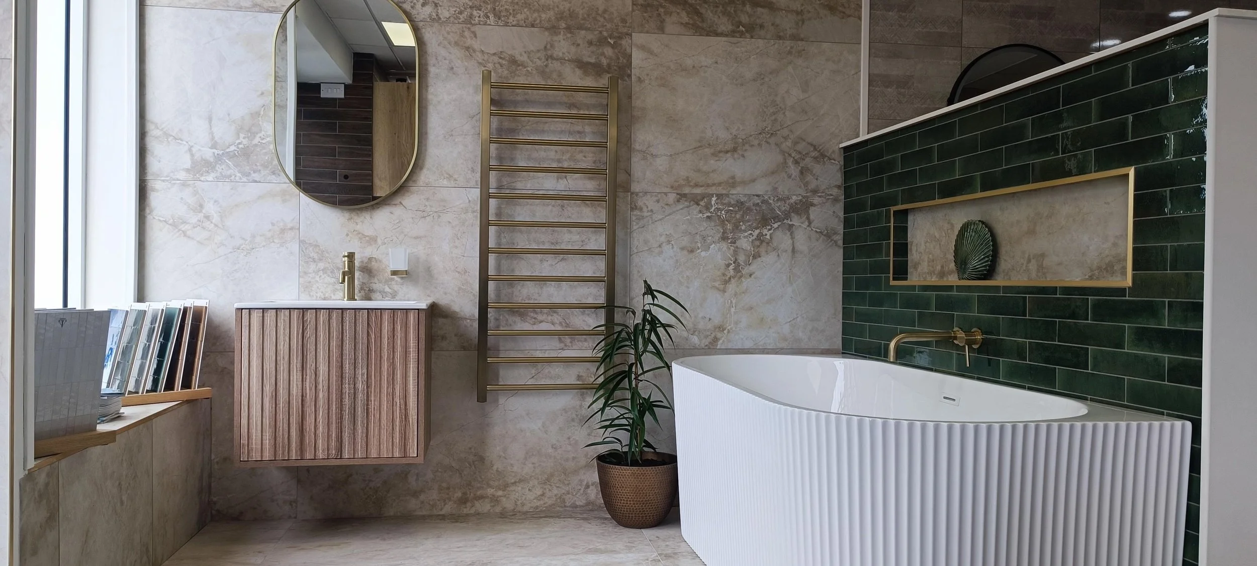 Modern bathroom with beige marble walls and flooring, a freestanding white bathtub, green tiled accent wall with a gold frame and decoration, a small vanity with a gold faucet, an oval mirror, a gold towel rack, a potted plant, and book stacks on a w