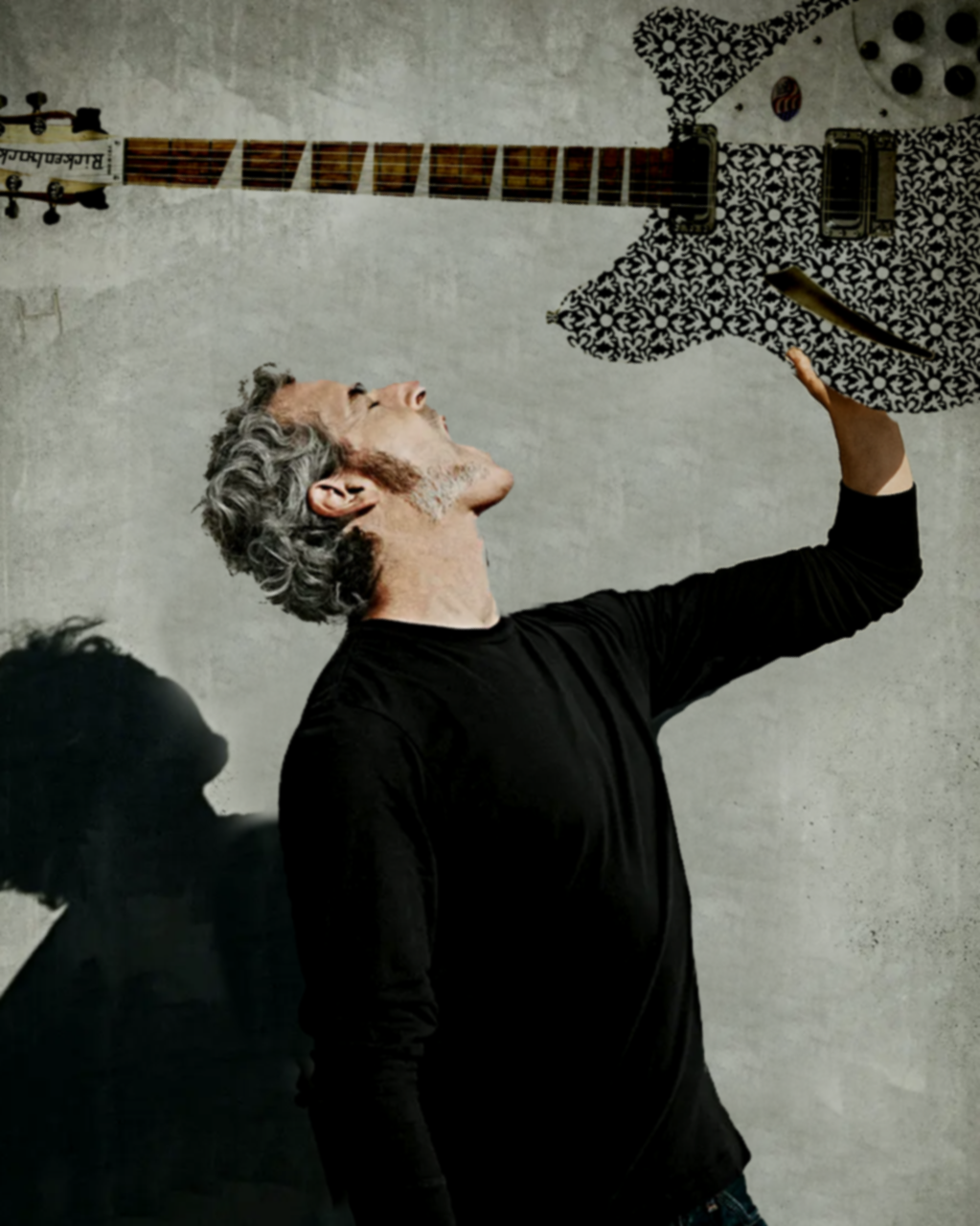 A man with gray hair and a beard, wearing a black long-sleeve shirt, is raising his right arm and looking up at a guitar hanging on a wall.