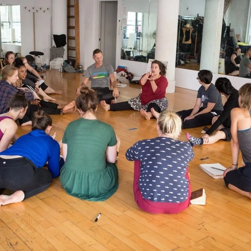 Group of people sitting on a wooden floor in a circle, engaging in a discussion or workshop, in a room with mirrors and large windows.