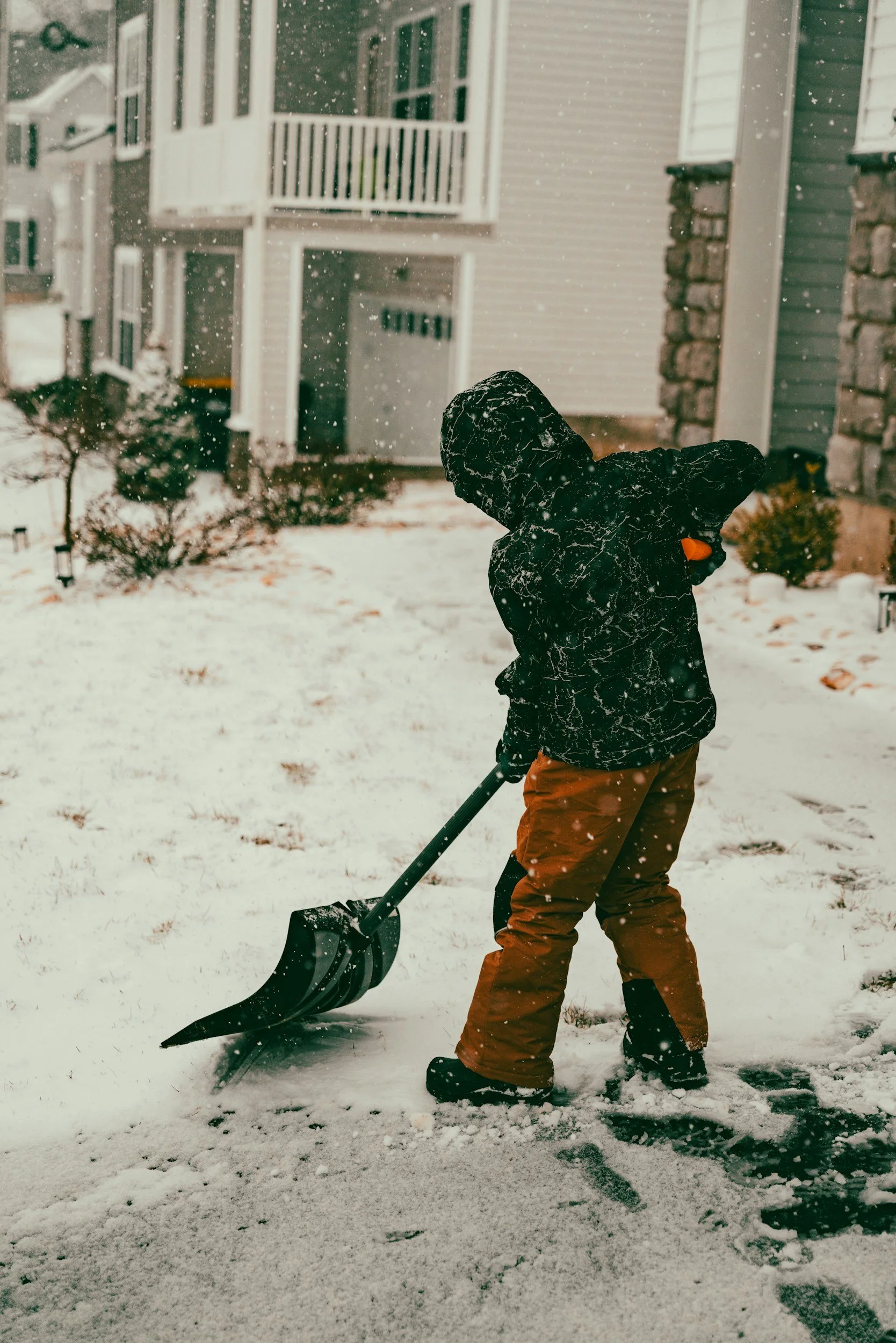 Worker shoveling snow in a residential backyard near a house with balconies and stone accents.