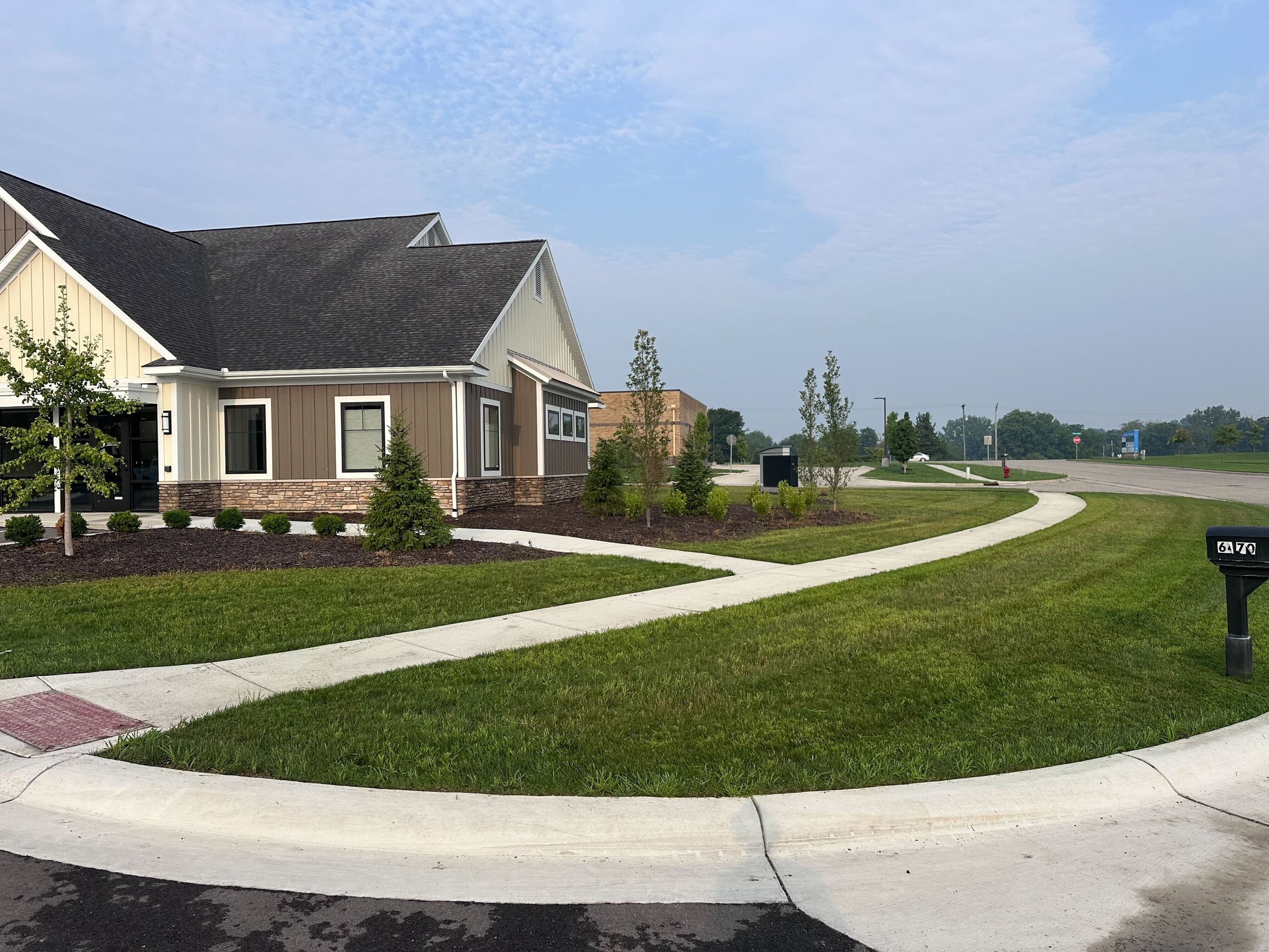 Residential neighborhood with a modern house, curved sidewalk, green lawns, trees, and a street on a cloudy day.