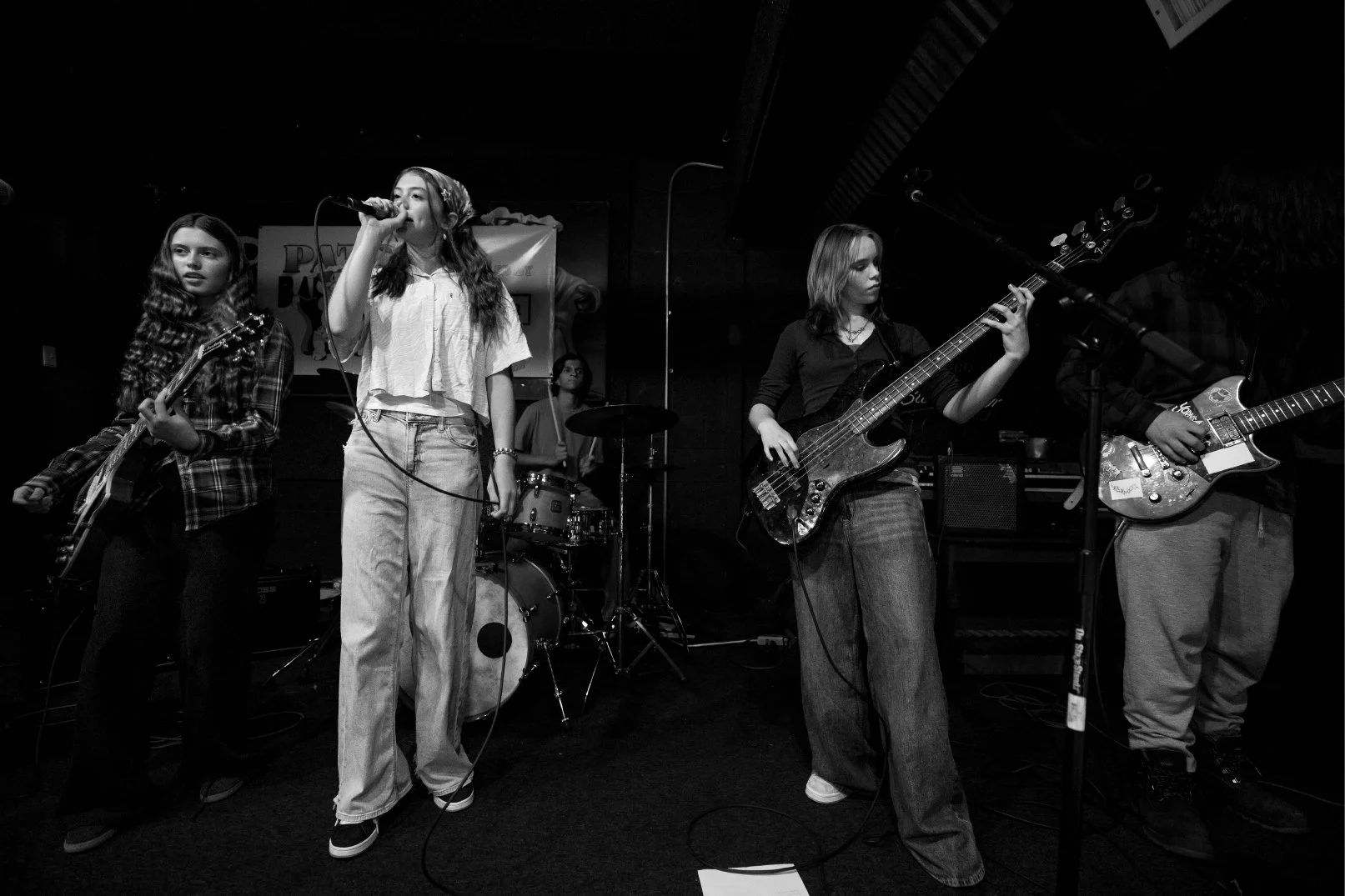 A band performs on a stage, including a singer with a microphone, a guitarist, a bassist, and a drummer, in a dark venue.