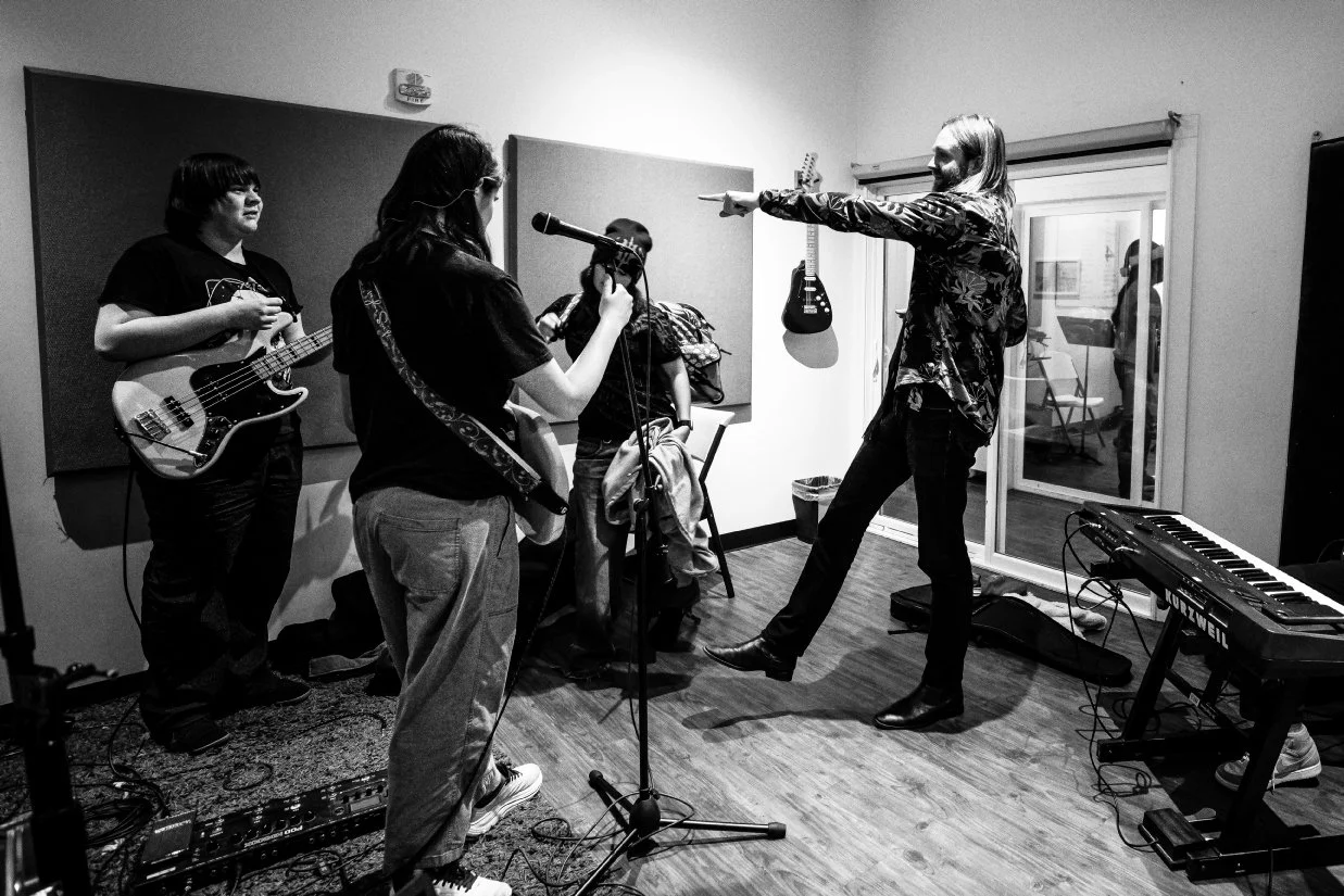 A music rehearsal or recording session in a studio with a man in a floral jacket leading, four young musicians with guitars, a keyboard, and microphones, and music equipment scattered on the floor.