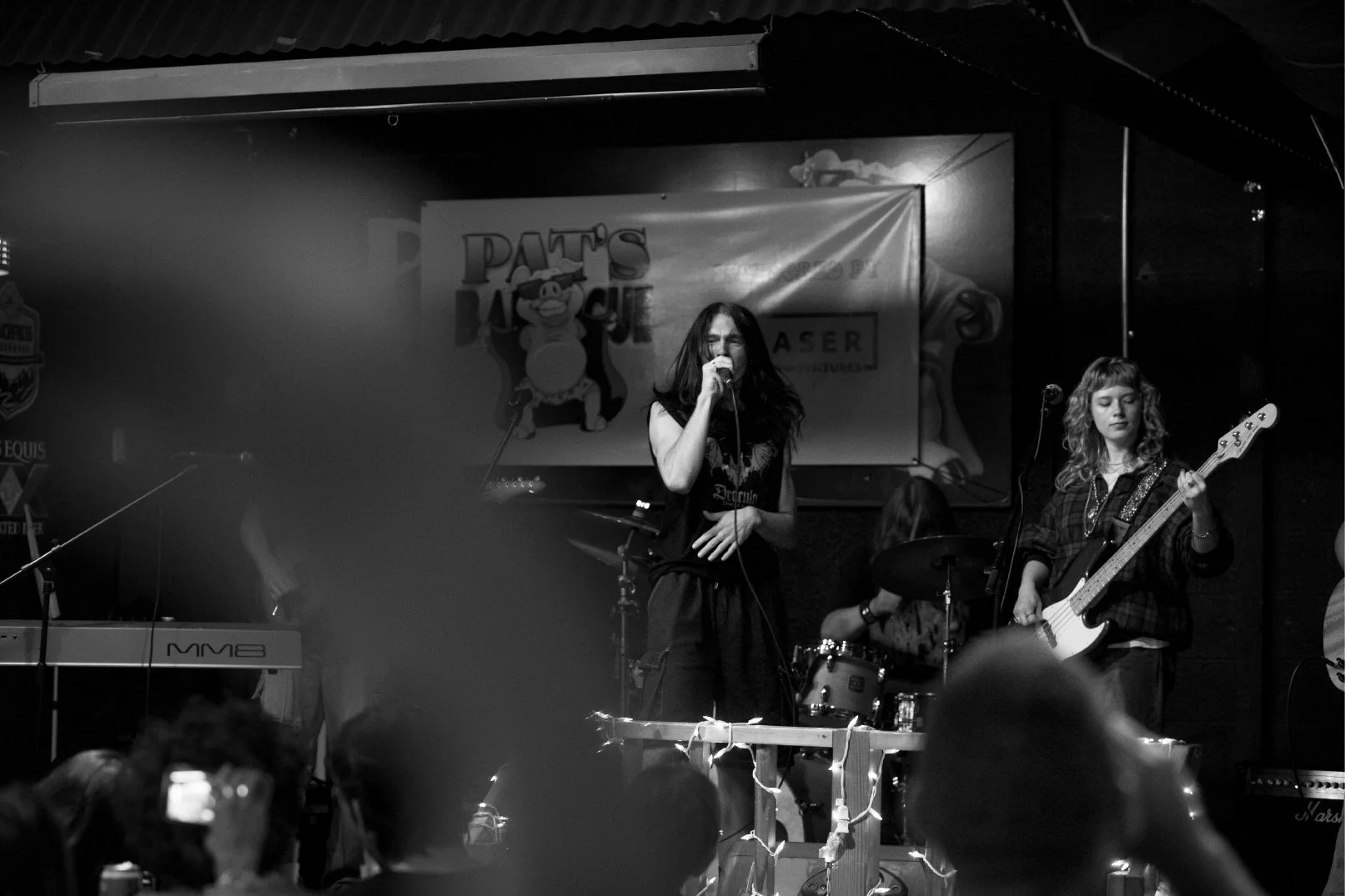 A black and white photo of a band performing on stage, with a female singer in the center, a female bass guitarist to her right, and a drummer in the background. Audience members are visible in the foreground.