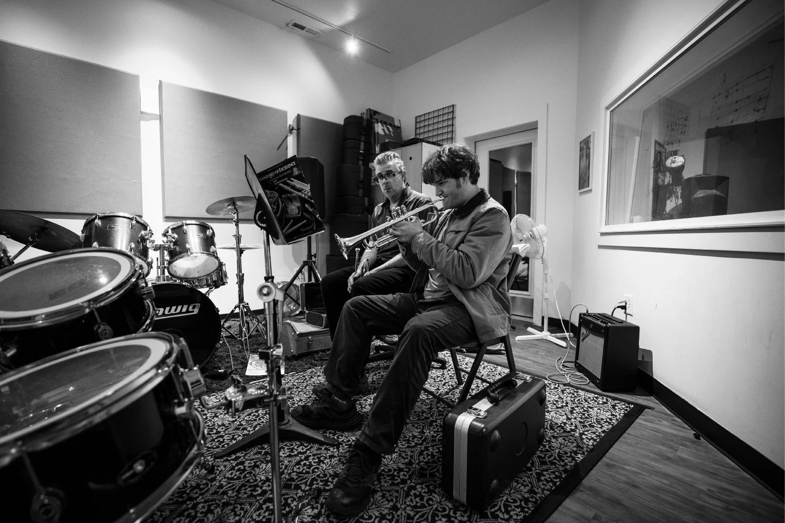 Two musicians, one playing trumpet and the other with a percussion instrument, in a rehearsal room with drums, music stand, amplifier, and various musical equipment.