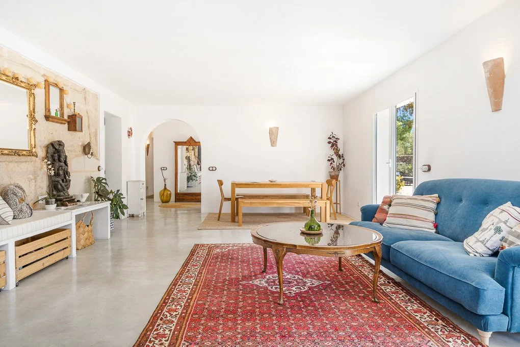 Living room with blue sofa, wooden coffee table, and red oriental rug, with a dining area and large windows.