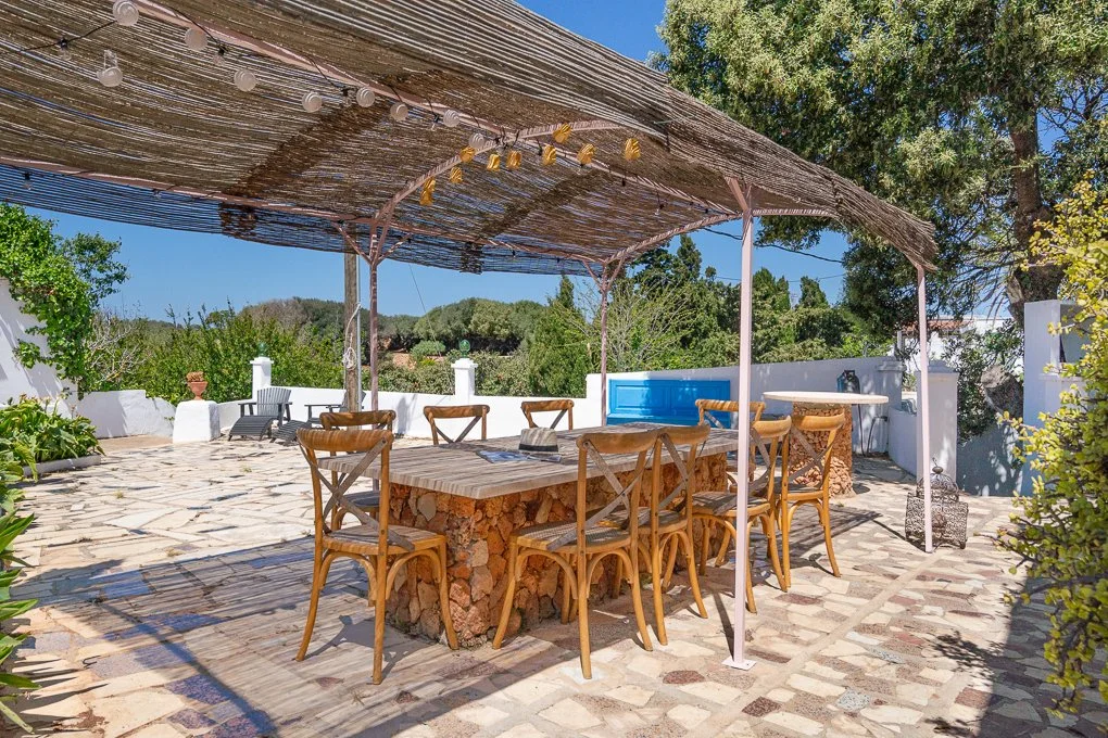Outdoor patio area with a wooden dining table and chairs under a shaded pergola, surrounded by trees and potted plants, with a blue pool in the background and clear blue sky.
