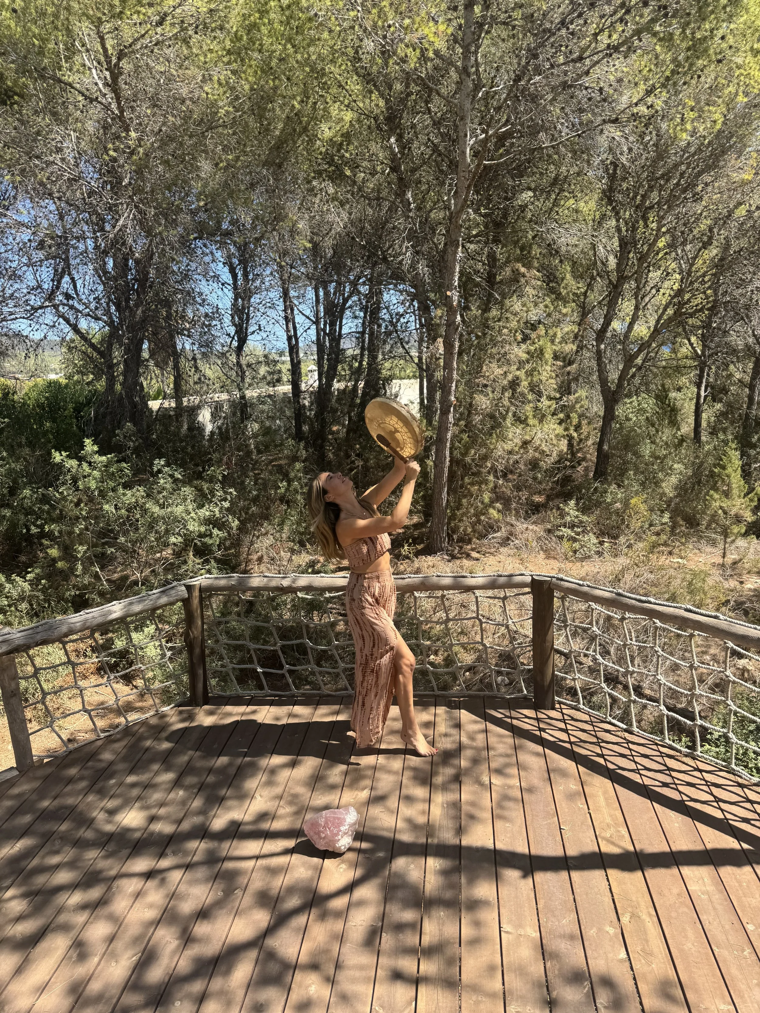 Woman standing on wooden deck surrounded by trees, holding a straw hat above her head and looking up, with a pink crystal on the deck.