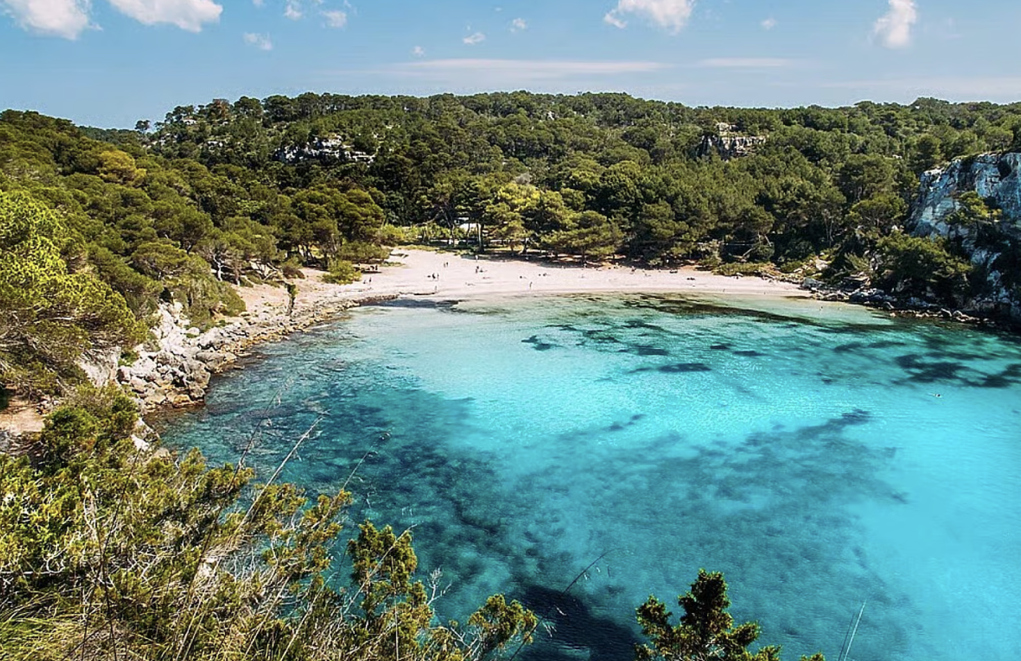 A secluded beach with turquoise water, surrounded by green trees and rocky cliffs under a partly cloudy sky.