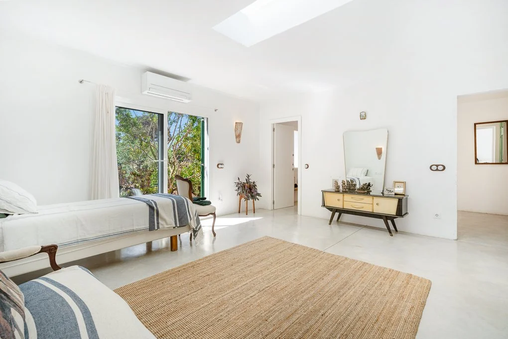 Bright, minimal bedroom with white walls, skylight, window with view of trees, and simple furniture including beds, a dresser with mirror, and a chair.