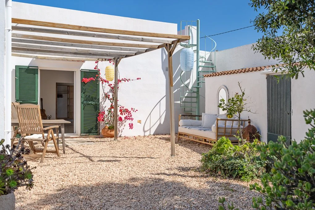 Small outdoor patio with gravel ground, wooden table and chairs, white sofa, potted plants, and a spiral staircase with hanging lanterns against white walls and blue sky.