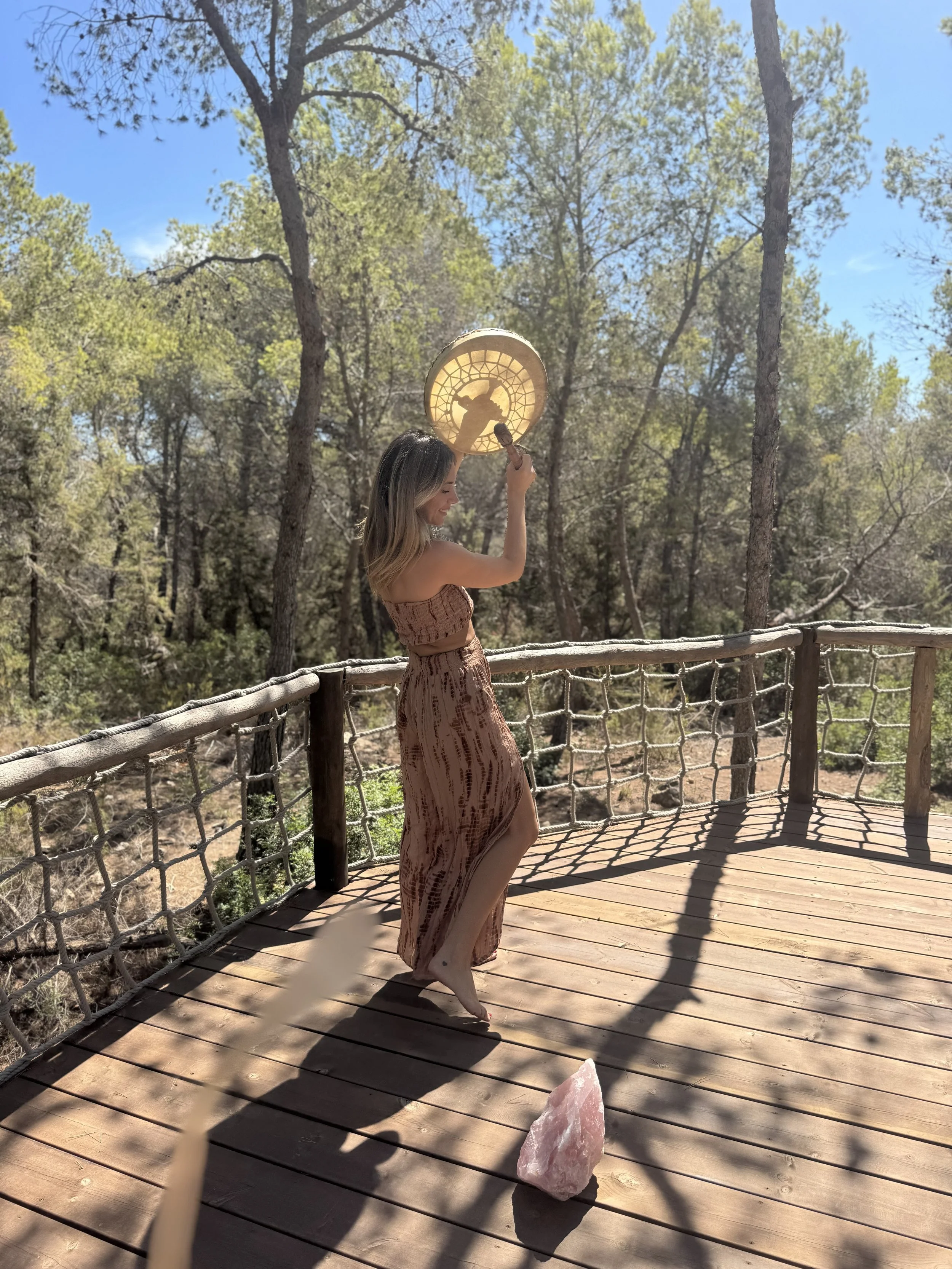 A woman in a patterned brown dress standing barefoot on a wooden deck surrounded by trees, holding a moon-shaped lantern with a silhouette of a horse against the countryside.