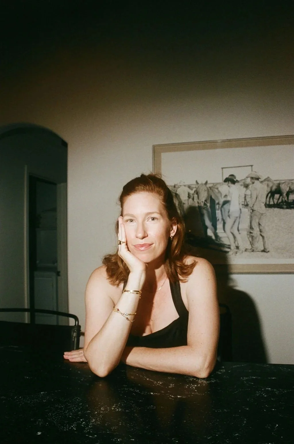 A woman with red hair, wearing a black top, sitting at a dark table, resting her head on her hand, with a framed drawing of a group of people on a beach in the background.