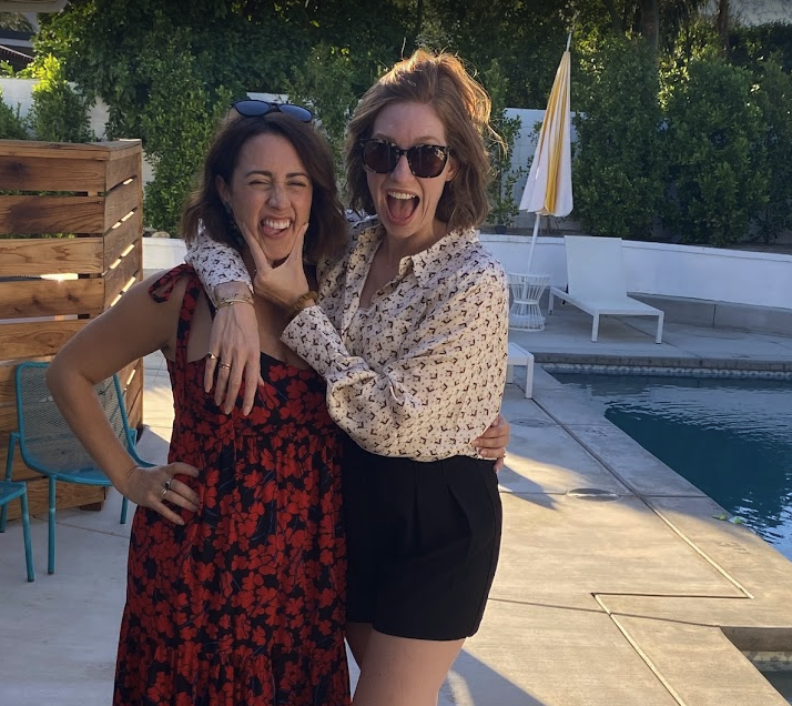 Two women smiling and laughing by a pool. One is wearing a red and black floral dress, and the other is wearing a patterned blouse and black shorts. They are outdoors with trees and poolside furniture in the background.