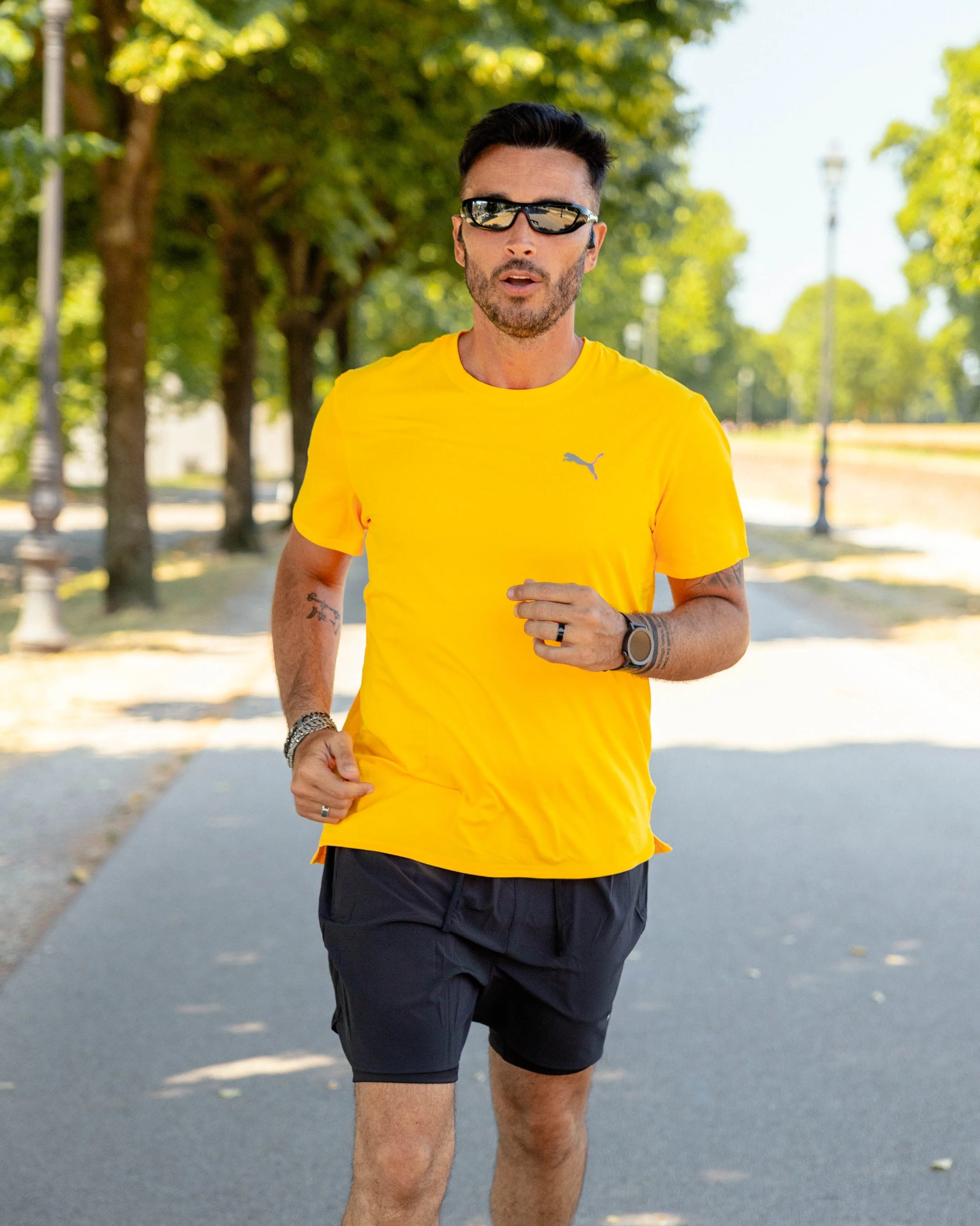 Uomo che corre in un parco soleggiato con maglietta gialla, pantaloncini neri e occhiali da sole.