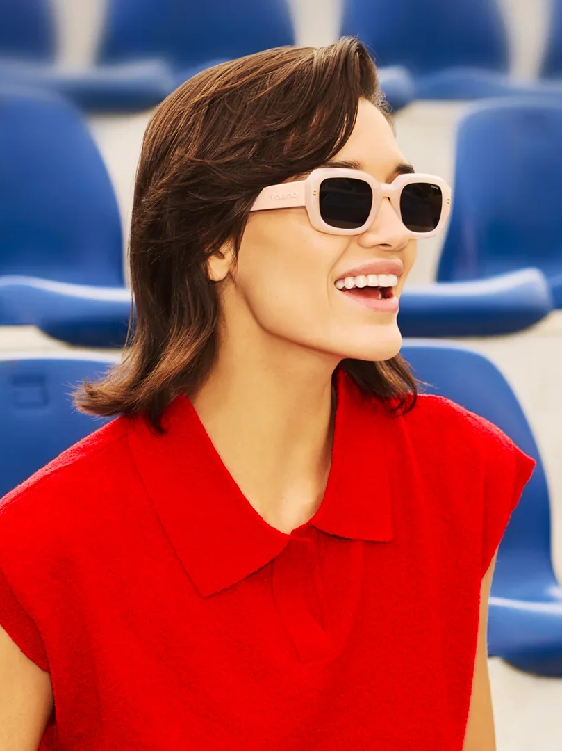 Una donna sorridente con capelli castani, indossa occhiali da sole bianchi e una maglia rossa, seduta in uno stadio con sedie blu.